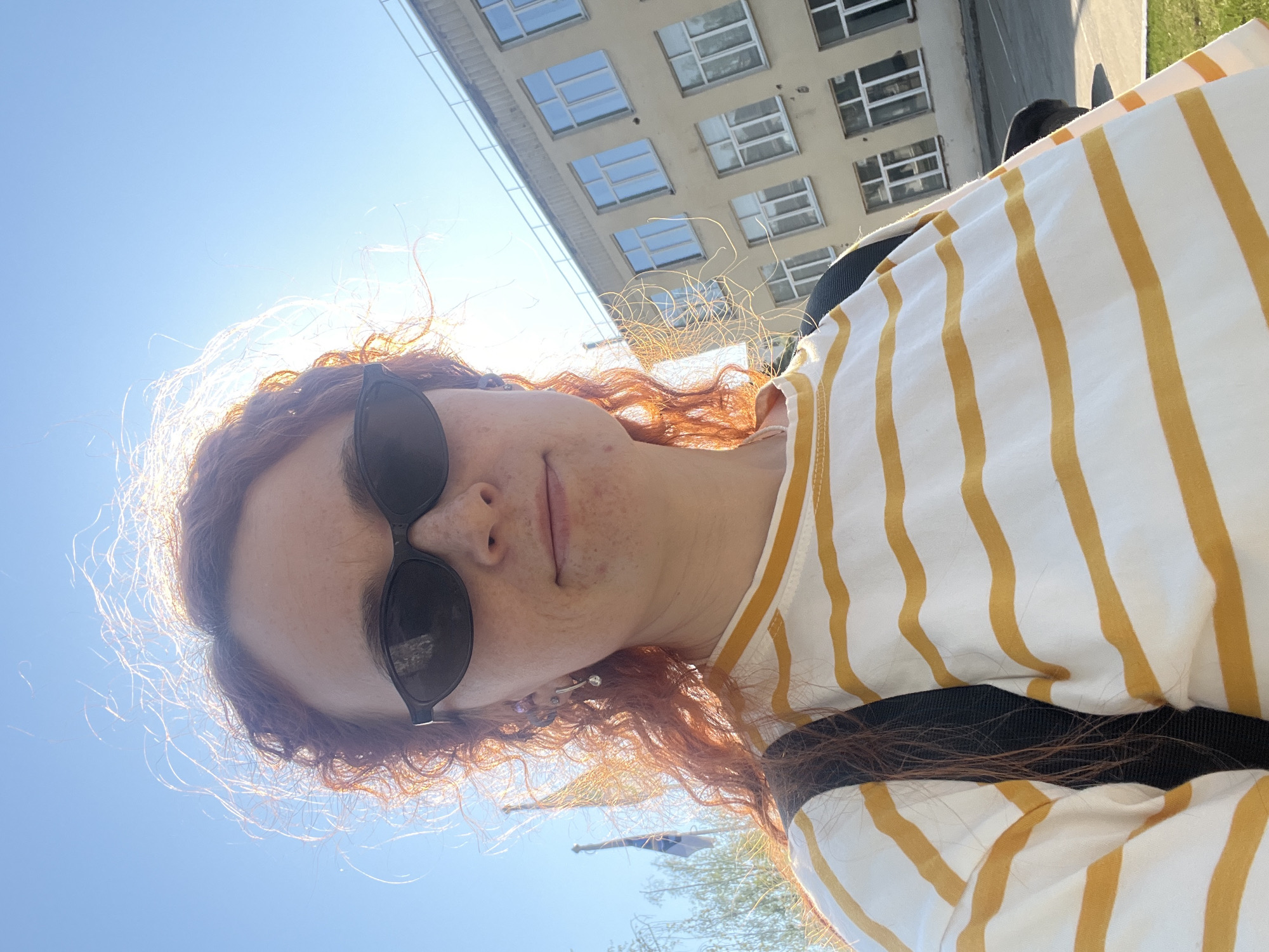 A selfie of a person with fair skin and red, curly hair wearing sunglasses and a white and yellow striped shirt is oriented upwards towards a building. The building, with many windows, is made of light-colored stone. The sky is visible behind the person and the building. The person is wearing a black backpack. A street and trees are barely visible in the background. The location may be Chaikovsky, Russia.