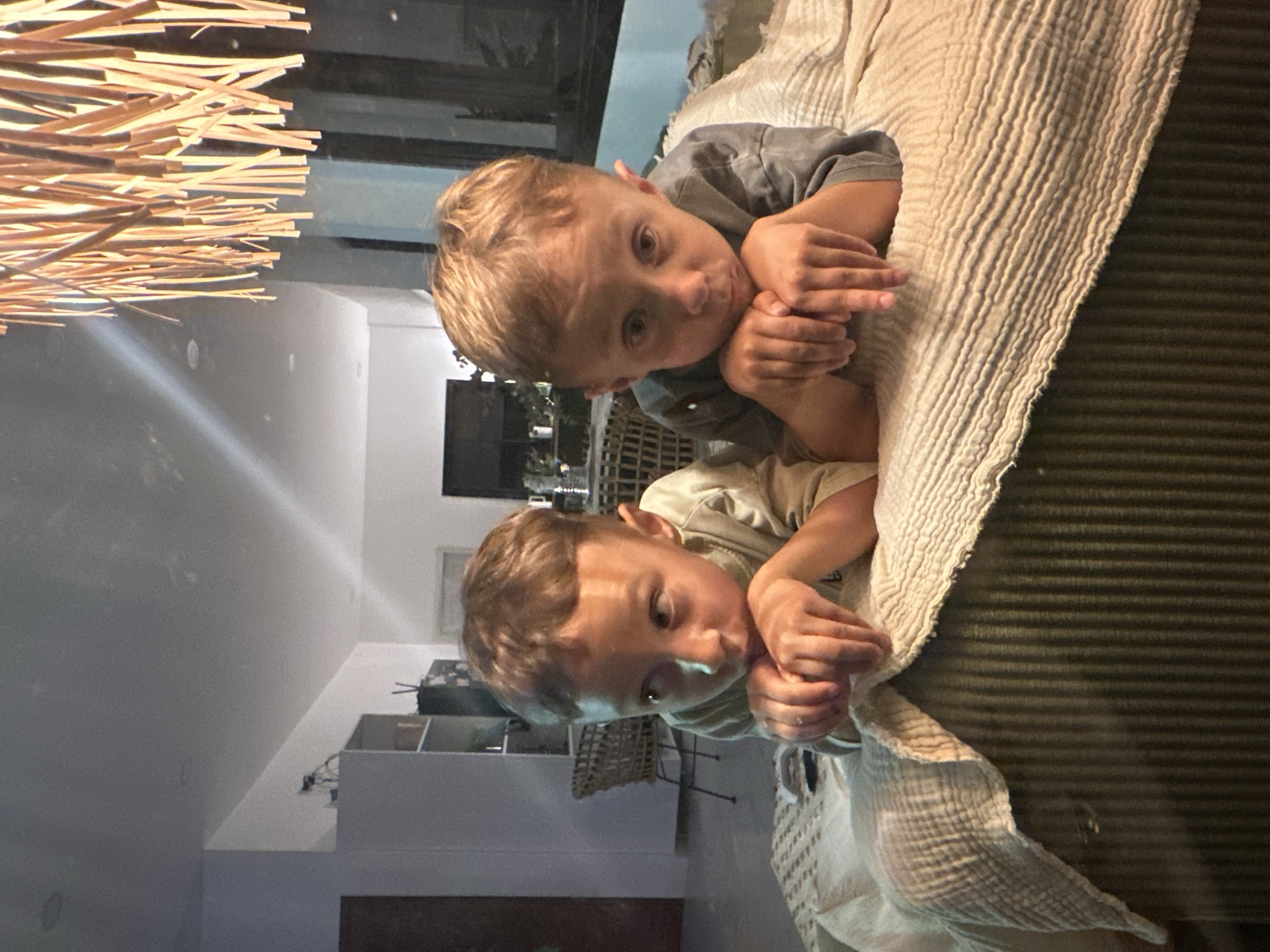 Two young boys are lying down on a textured, light-colored blanket and a dark green, ribbed surface, possibly a couch. The boy in the foreground, with blonde hair, is looking down and resting his chin on his hands. He is wearing a light-colored t-shirt. Behind him, another boy, also with blonde hair, is looking towards the camera. He is wearing a dark grey t-shirt and has his hands clasped in front of him. The lighting suggests it is indoors, possibly in the evening, with artificial light creating soft shadows. In the background, a white wall is visible, along with shelves and what appears to be a woven basket. To the left of the frame, a decorative element made of thin, light-colored wooden sticks is hanging, creating a patterned overlay on the image. The overall mood appears calm and relaxed. No text is visible in the image.