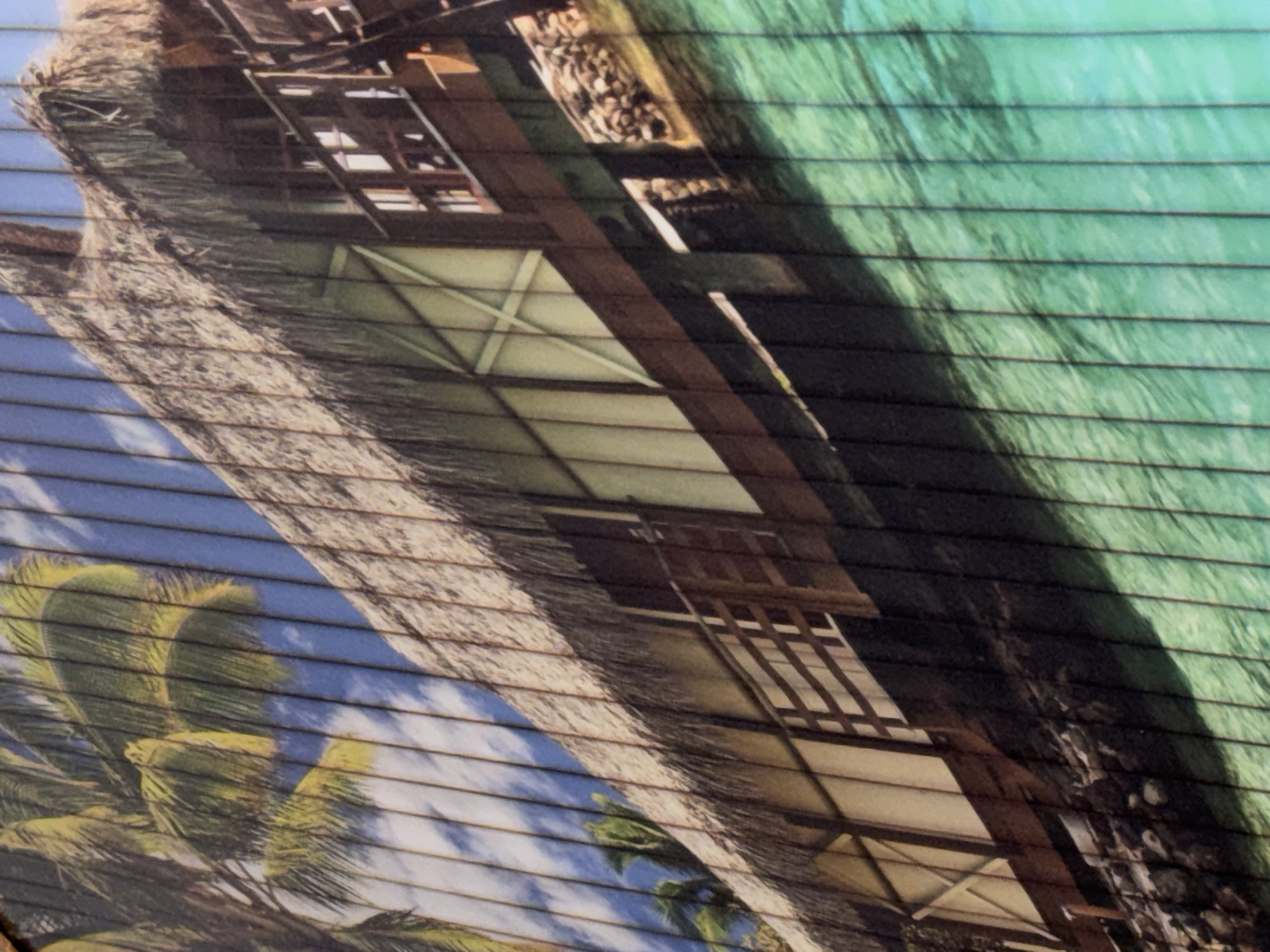 The image shows a structure, possibly a resort building, with a thatched roof. The building has wooden supports and what appear to be windows or open-air spaces. There are palm trees visible in the bottom left corner, and the background on the right side of the structure has a greenish-blue color, possibly representing water. The overall scene suggests a tropical or island location. The image shows a 3D printed representation of an island and the photo appears to be a picture of the 3D printing layers. It is unlikely to be located in Perm, Russia.