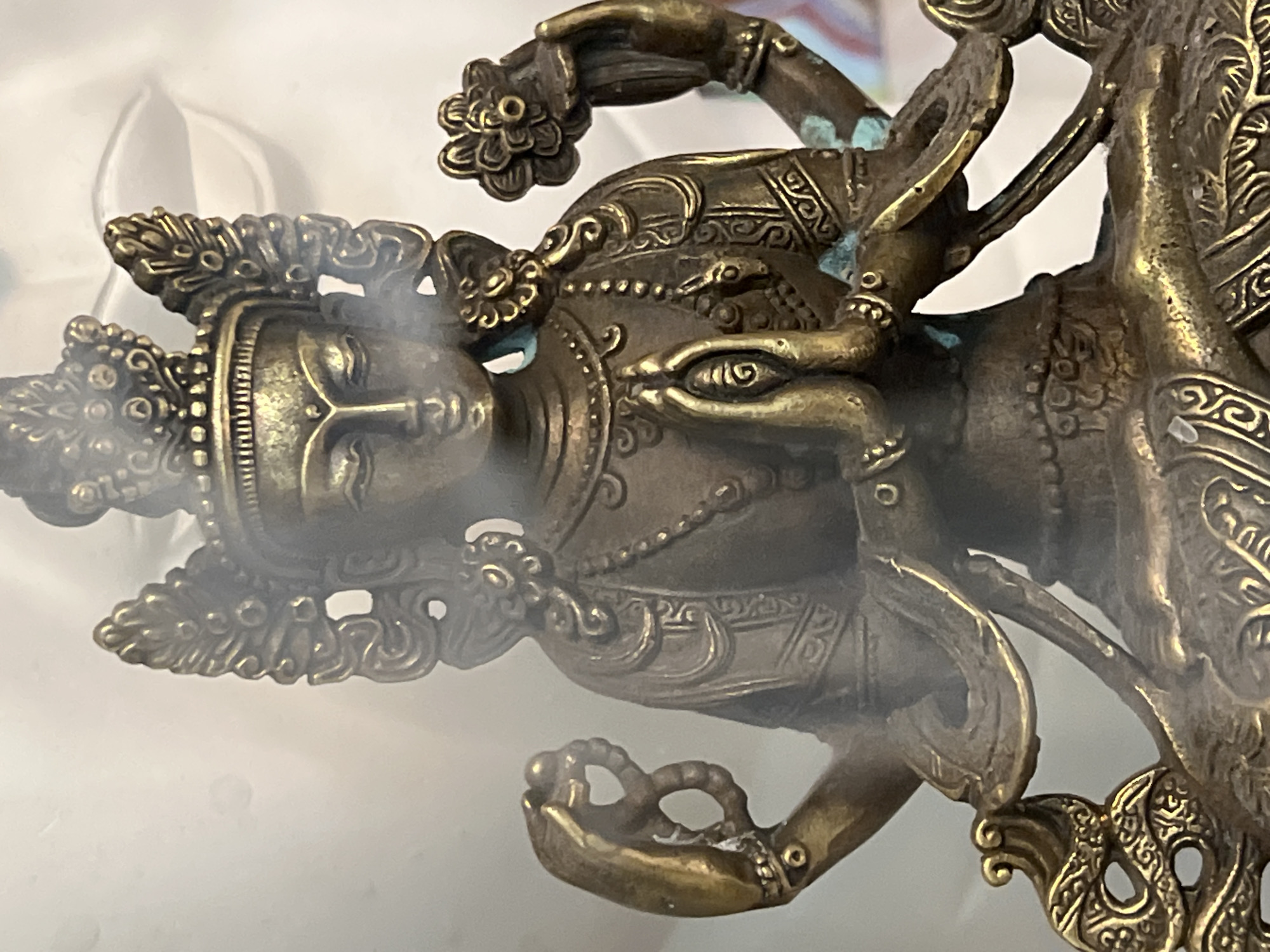The image shows a close-up of a bronze statue of a multi-armed deity, possibly a Buddhist or Hindu figure. The statue is detailed, with intricate carvings on its crown, jewelry, and clothing. Smoke or mist is rising from around the deity's head and upper body. The statue is placed against a blurred background, which appears to be an indoor setting with a white or light-colored surface. There is no indication of the city of Petrozavodsk, Russia, or any other specific location within the image.