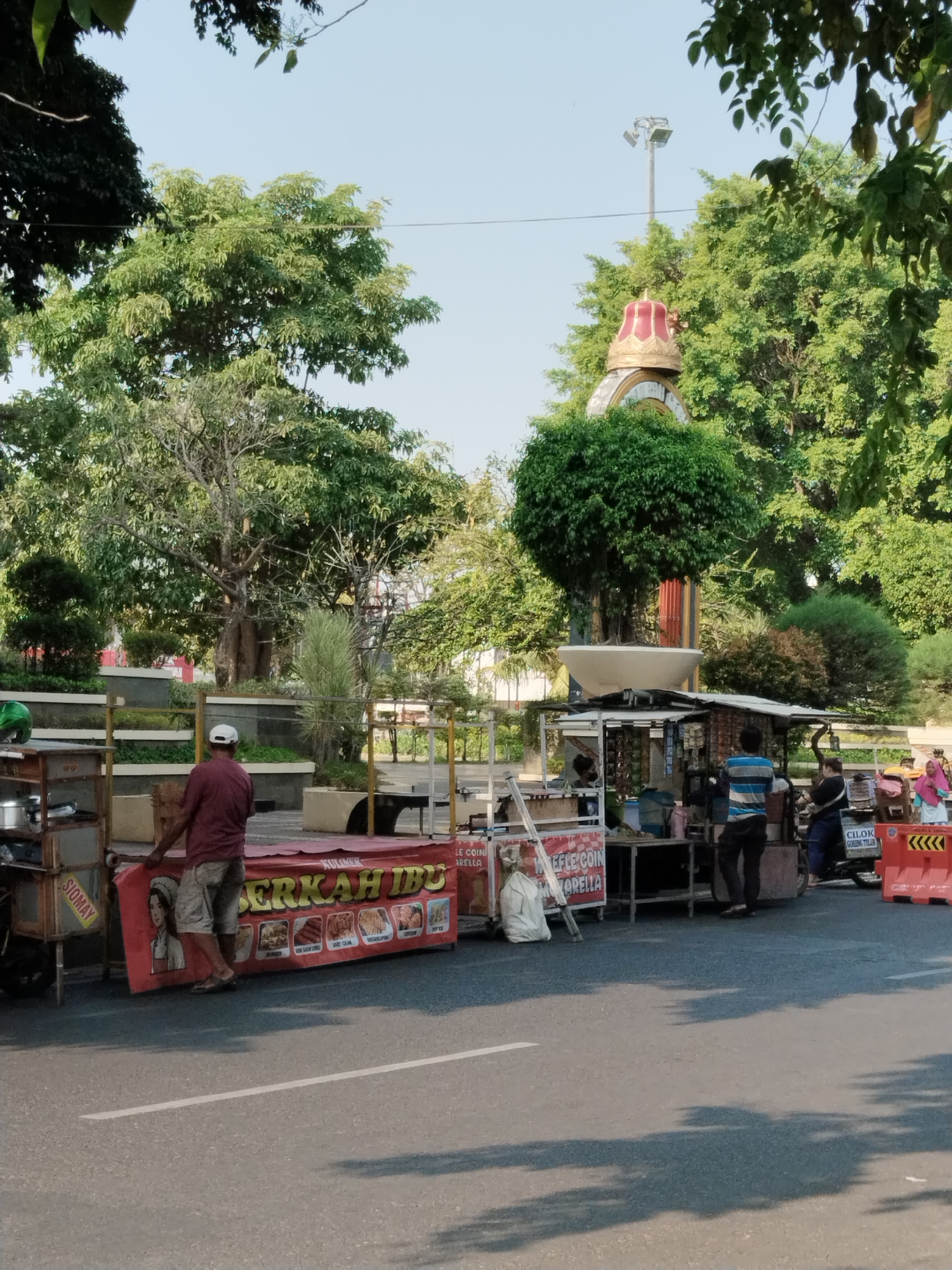 The image depicts a street scene in Kecamatan Pati, Indonesia, on a sunny day with a clear blue sky. Several food stalls are set up along the roadside, with awnings and banners displaying food items. In the foreground, a man in a maroon shirt and beige shorts stands near a food cart, holding a red banner. To his right, another stall is visible with people behind the counter. Further down the street, more stalls and individuals are present, including a woman in a pink hijab. In the background, lush green trees and a monument topped with a red and gold crown are prominent. The street appears to be paved with asphalt, and there are some white road markings visible. The overall atmosphere suggests a bustling local market or street food area.