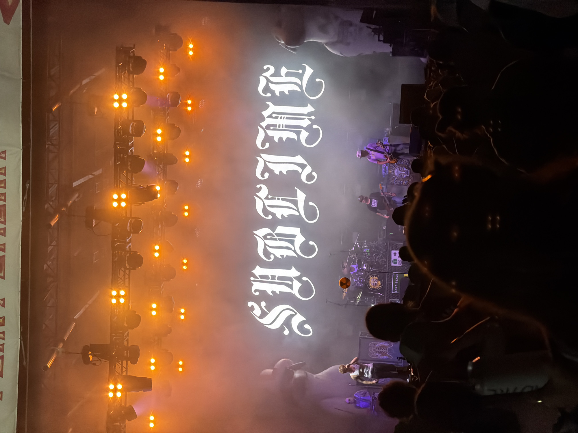 This is a photograph of a live music performance by the band Sublime at the Oceanfront Stage in Virginia Beach, Virginia. The band's name, "SUBLIME," is displayed in large, gothic-style white lettering on a dark screen behind the performers. The stage is illuminated by numerous bright orange lights, creating a hazy, atmospheric effect. In the foreground, a crowd of concert-goers is visible, with their silhouettes partially obscuring the view of the stage. The lighting suggests the performance is taking place at night or in a dimly lit venue. The overall atmosphere is energetic and typical of an outdoor concert event.