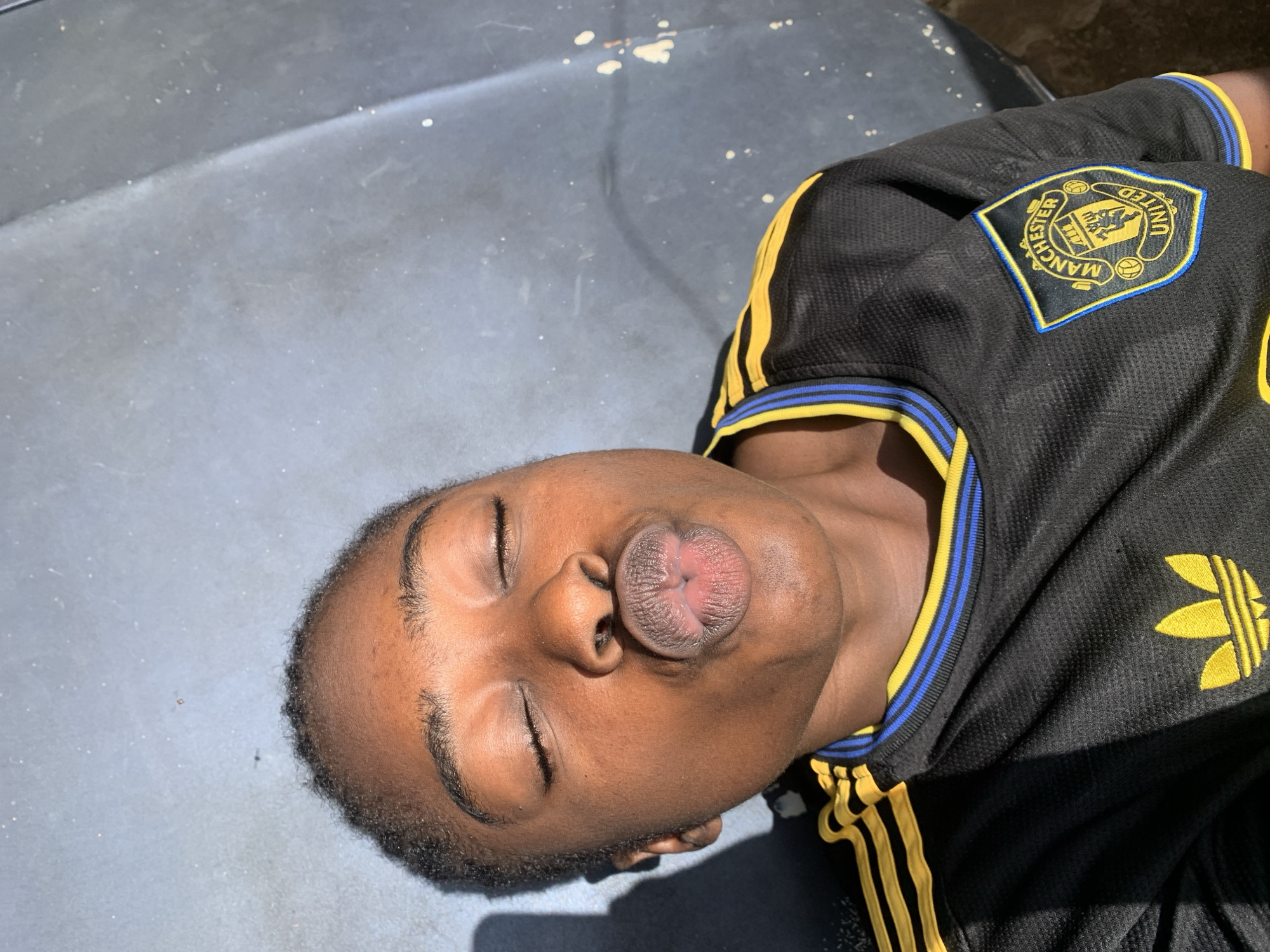 A close-up shot shows a person with their eyes closed and lips puckered, appearing to blow a kiss. They are wearing a black Manchester United jersey with yellow and blue trim. The person is lying down on a dark, textured surface, possibly the hood of a vehicle, outdoors. The lighting suggests it is daytime, with harsh shadows visible on the left side of the image. The background is a gray, somewhat speckled surface that could be a road or a weathered exterior. The image appears to have been taken in Rafin Par, Nigeria, given the context.