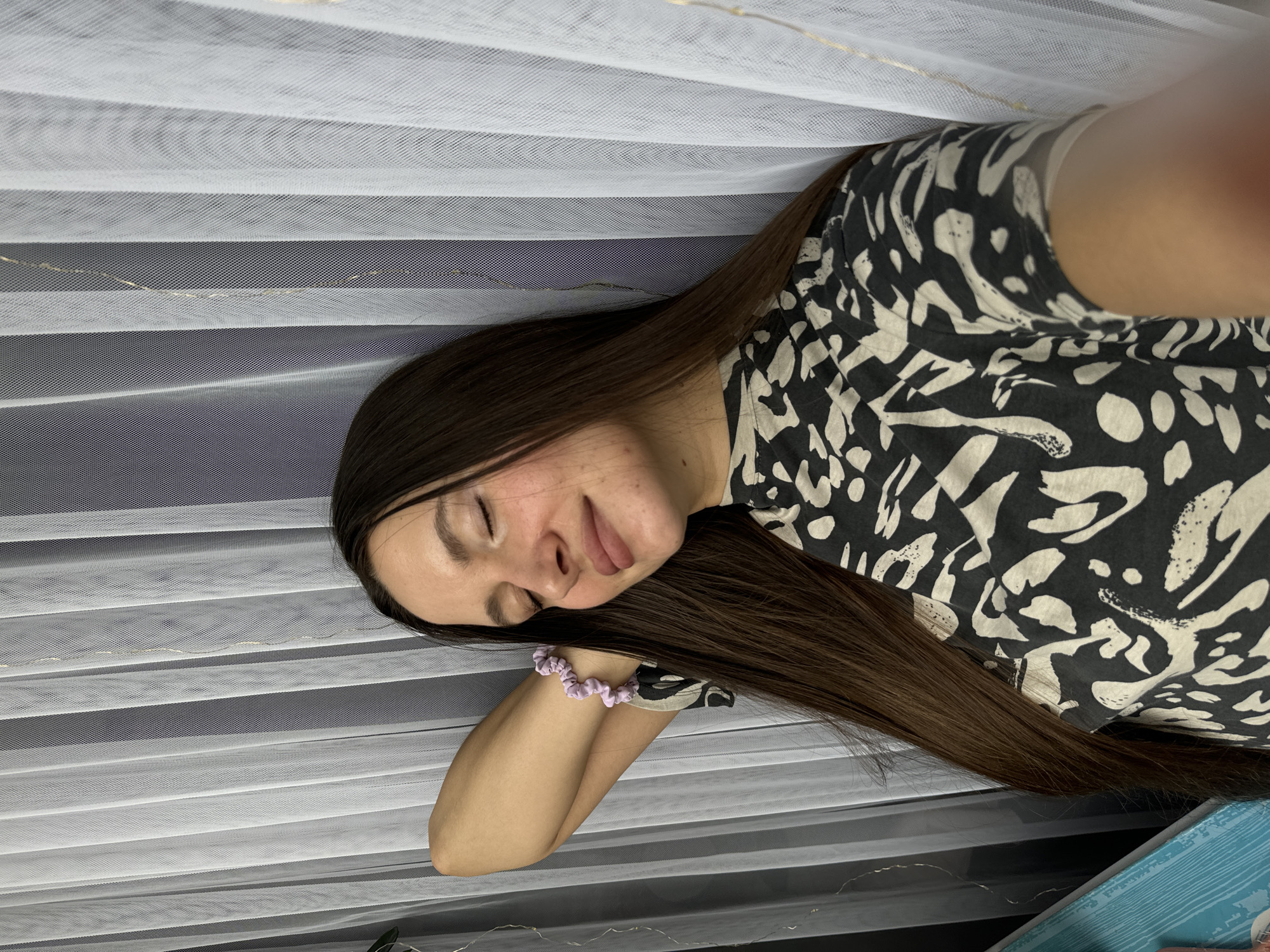 A young woman with long, dark brown hair is captured in a close-up indoor shot, appearing to be resting or asleep. Her eyes are gently closed, and her face holds a peaceful expression. She is wearing a black and off-white patterned top with an abstract or animal print. A light purple, scrunchie-style bracelet adorns her right wrist, which is bent, suggesting her arm is supporting her head.
The background is dominated by a white, translucent pleated fabric, likely a curtain or blind, forming distinct horizontal stripes. Through this sheer material, a darker, possibly gray or blue, background is visible. A faint string of small, golden lights (fairy lights) can be seen delicately threaded within or behind the pleated fabric, adding a subtle glow. In the lower right corner of the frame, a segment of a textured blue object, possibly a book or a decorative box, is partially visible. The overall atmosphere of the image is serene and tranquil, characteristic of a restful moment in a cozy indoor setting in Akulovo (Gp Kubinka), Russia. No text is visible in the image.