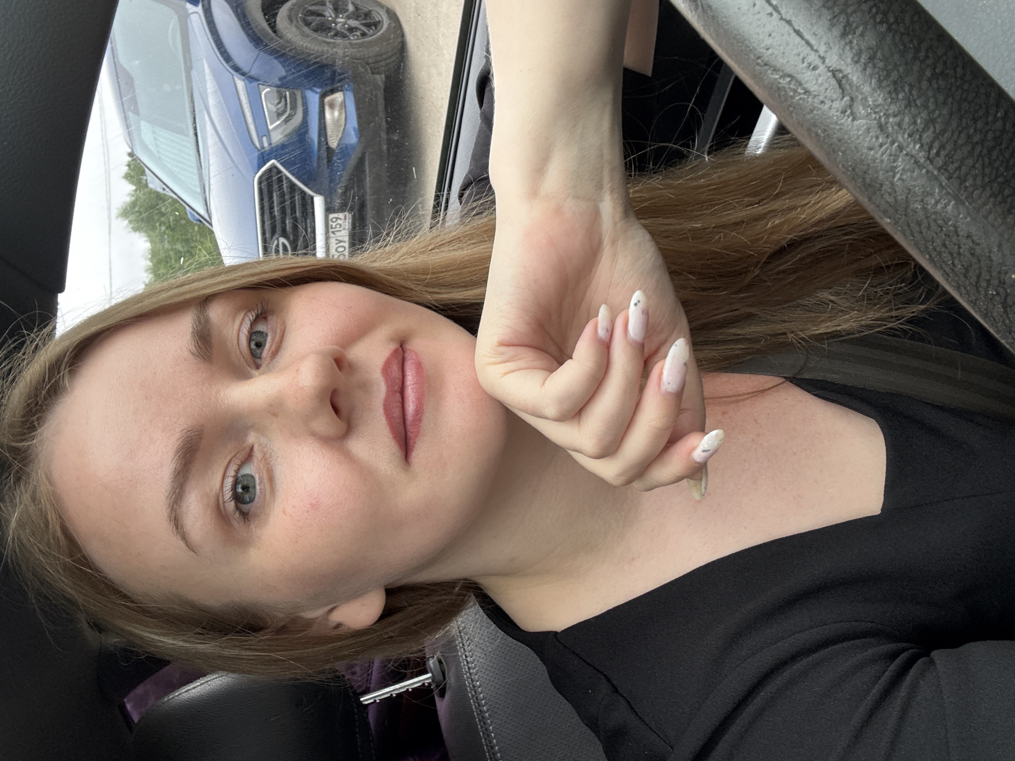 A woman with blonde hair is sitting in the driver's seat of a car, facing the camera. She is wearing a black shirt and has her left hand resting on her chin. Her nails are painted with a light pink polish and decorated with small white and silver accents. A blue SUV is visible through the car's windshield, and a license plate with the inscription "SOY 159" is partially visible on the SUV. The scene appears to be in an outdoor setting, possibly a parking lot, with trees and a cloudy sky in the background. The image was taken in the city of Chaikovsky, Russia.