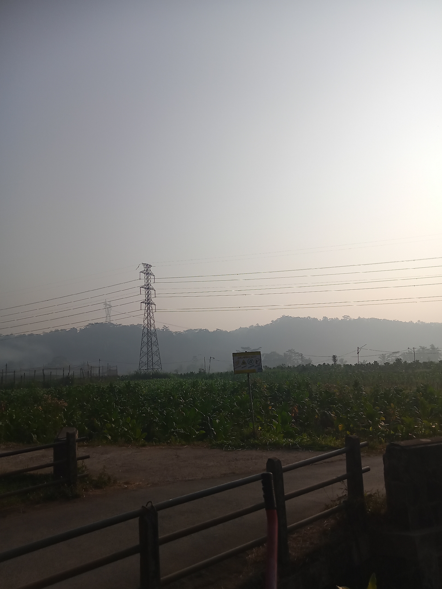 A wide shot captures a rural landscape on a hazy morning, likely in Sruwen, Indonesia. The foreground features a dark metal railing with wooden posts, separating a dirt path from what appears to be a fenced-off area with dense green foliage, possibly crops like tobacco or corn. Beyond this, a dirt road curves to the right. In the mid-ground, a large metal electricity pylon stands prominently, with several power lines stretching horizontally across the frame. Another, smaller pylon is visible in the distance, partially obscured by mist. A signpost with multiple warning symbols is situated to the left of the main pylon. The background consists of rolling hills covered in lush vegetation, shrouded in a light mist, with a bright, hazy sky above. The overall atmosphere is serene and agricultural, characteristic of a rural setting.