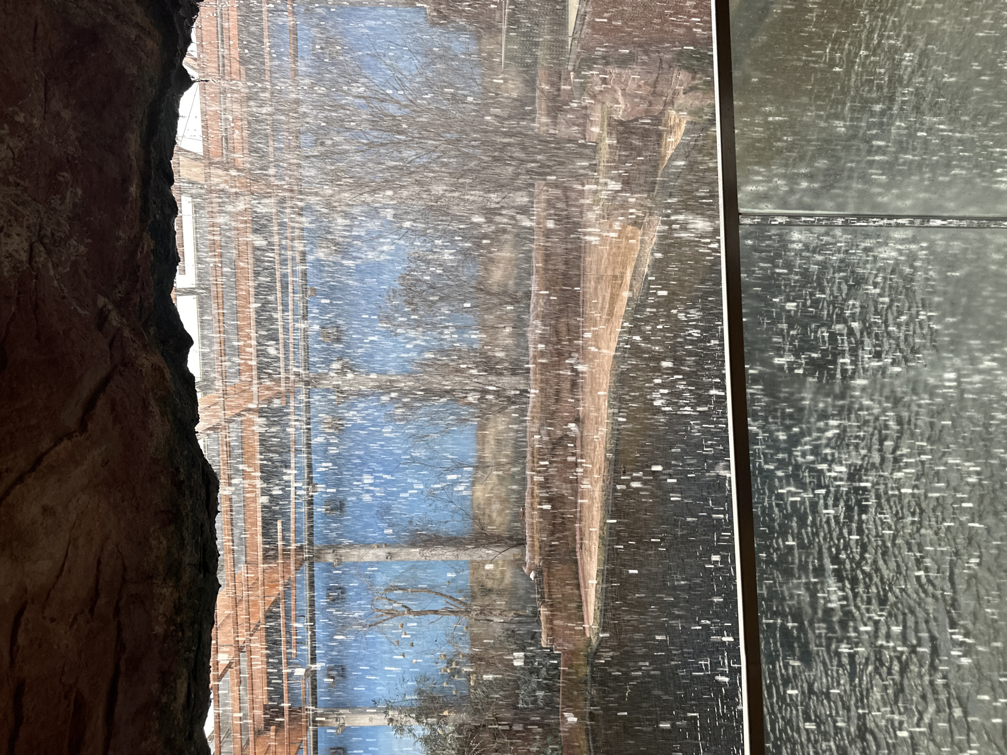 The image shows a view from a cave or rocky alcove onto a scene in Wrocław, Poland. Outside the alcove, it appears to be snowing, or the view is distorted by moisture. In the distance, there is a brick building with blue sections. Bare trees are also visible. To the right, there are reflective or glossy textured panels or tiles.
