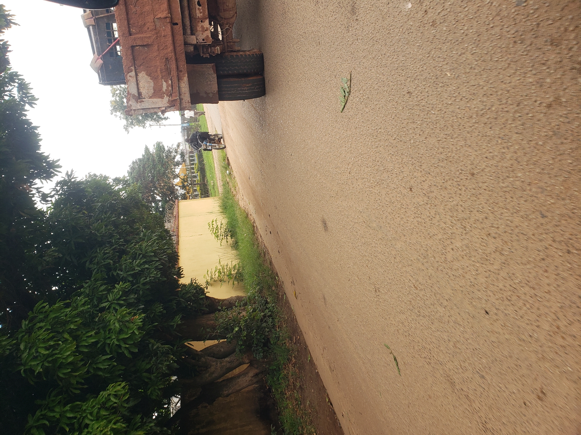 The image depicts a street scene in Gura Top, Nigeria. In the foreground, a paved road occupies the majority of the frame, with a rough, textured surface. A few scattered leaves lie on the road. To the left, a yellow building with a textured wall and a dark, rough stone wall is partially visible, lined with lush green foliage and the trunks of large trees. Beyond the building, a metal fence with barbed wire runs along the property line.
In the middle ground, a person is riding a motorcycle on the road, heading away from the viewer. The rider is wearing dark clothing and a helmet. Further down the road, another person is walking. To the right, the rear section of a large, rusty dump truck is parked. The truck's tires are visible, and its cargo bed is filled with reddish-brown material, possibly soil or aggregate. A red shovel is resting on the edge of the truck bed. The background shows a few more trees and a bright, overcast sky. The overall impression is that of a rural or semi-urban street during the daytime.