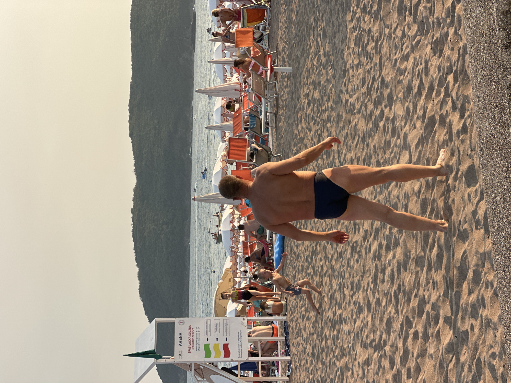 This image captures a lively beach scene in Boreti, Montenegro. In the foreground, a man in dark swim trunks stands facing away from the camera, his left arm slightly extended as if in preparation for an activity. To his left, a young boy in dark shorts runs across the sand, looking towards the man. The beach is populated with many people enjoying the day. Beach chairs and umbrellas are set up, with some individuals lounging and others walking around. In the background, the calm sea is dotted with boats, and a lush green hillside forms a picturesque backdrop. A lifeguard stand with flags and information signage is visible on the left side of the image. The overall atmosphere is one of recreation and relaxation on a sunny day.