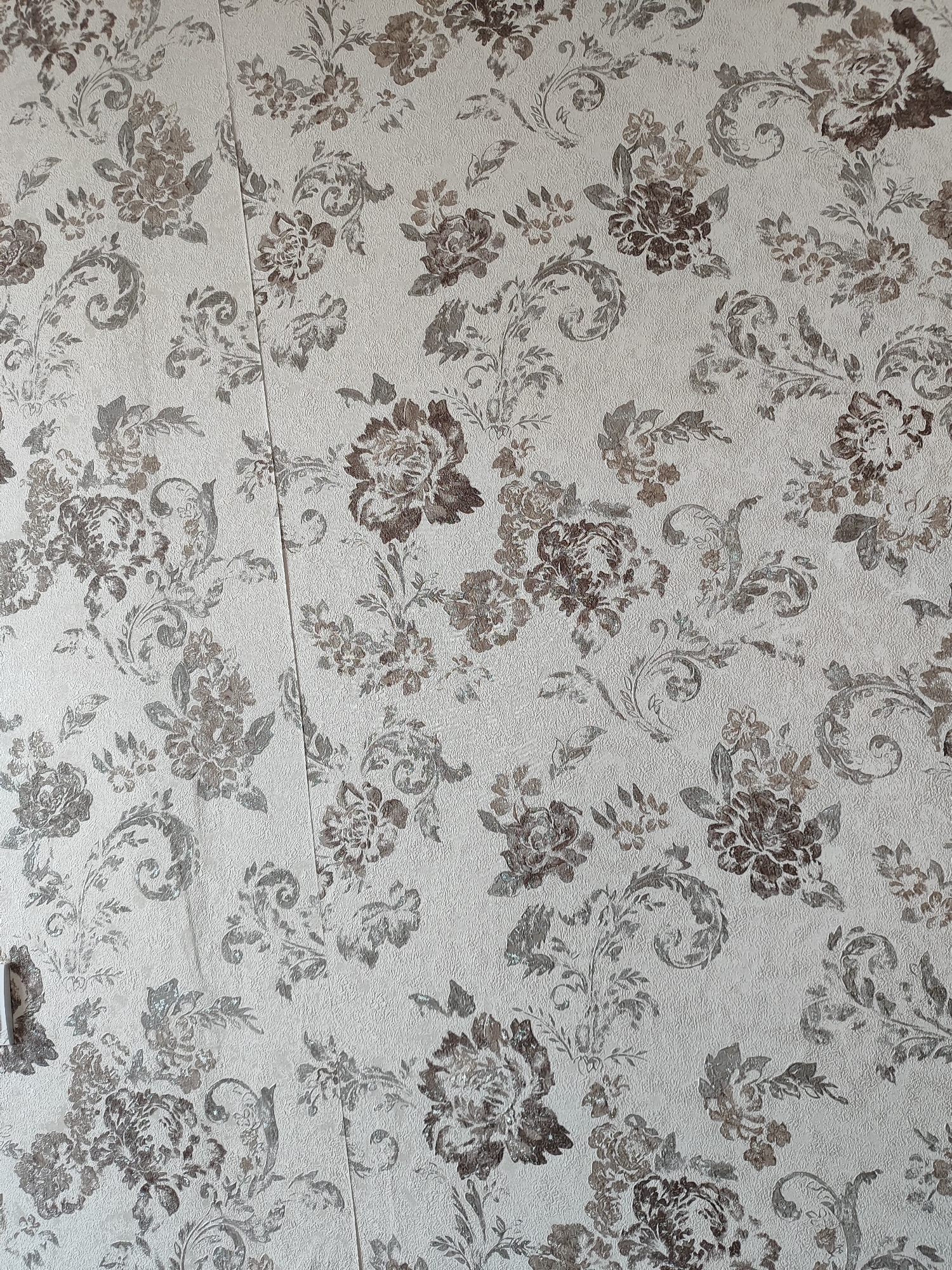 The image shows a close-up of floral wallpaper. The wallpaper has a textured, off-white background with a repeating pattern of large, dark brown and muted grey flowers and scrolling vines. The design is dense and appears to be printed or embossed onto the wallpaper. There is a visible seam running vertically down the center of the image, indicating the wallpaper is applied to a wall. The lighting is even, suggesting an indoor environment. There are no people, animals, or other objects visible in the frame. The image does not provide any information about the location in the city of Marusino, Russia.