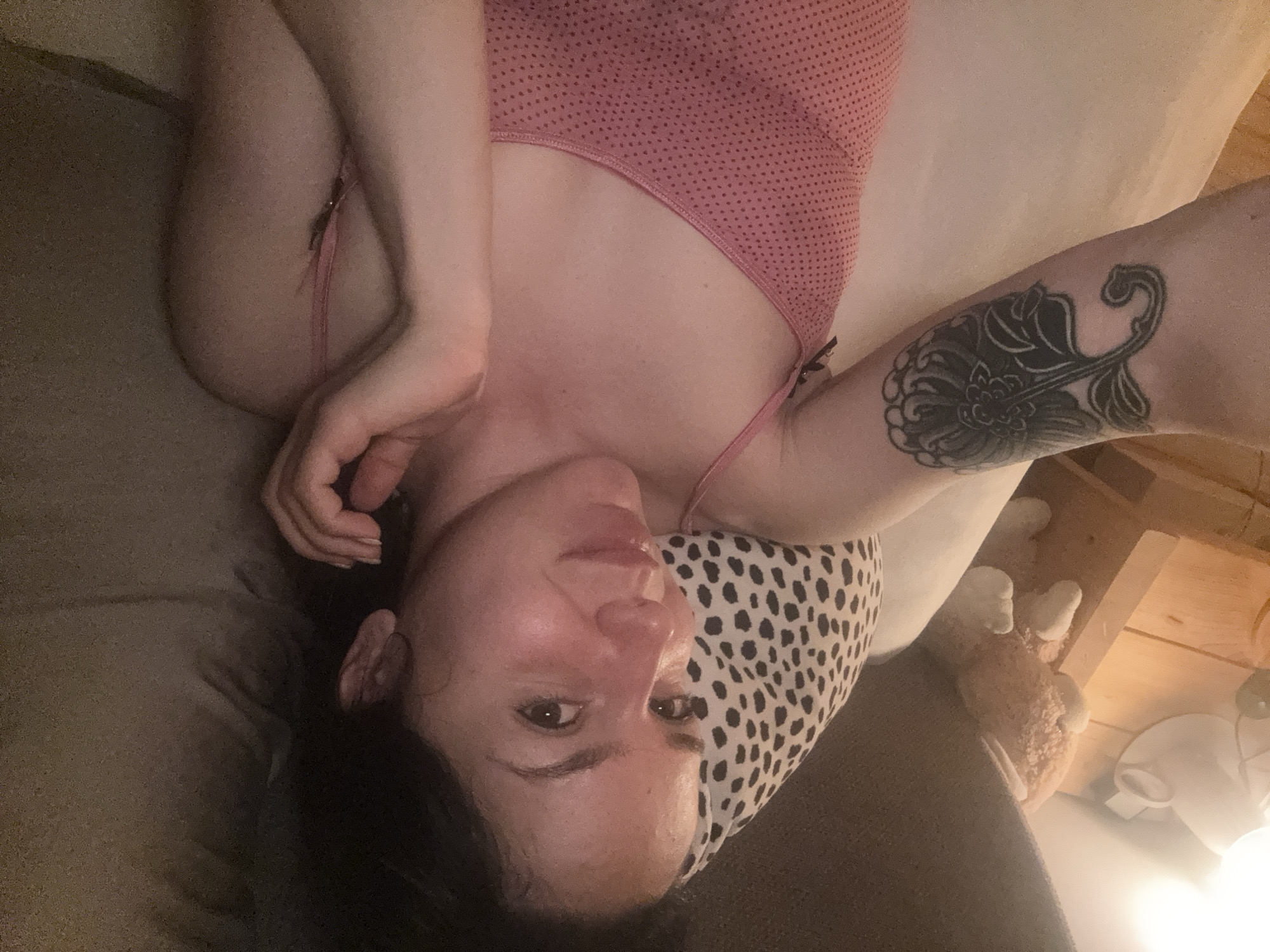 An adult person, appearing to be female, is depicted lying on her back, looking directly into the camera with a gentle smile. The image is oriented such that her head is towards the bottom of the frame. She has dark hair and fair skin. She is wearing a pink garment, possibly lingerie or a bodysuit, with a dotted pattern on the upper part and thin straps. A prominent black tattoo, featuring an elaborate floral or abstract design, is visible on her right forearm (viewer's left). Her left arm is bent, with her hand near her chest, while her right arm extends upwards.
She is resting on a bed or sofa, with a white pillow adorned with black spots beneath her head. The background features light-colored bedding and hints of wooden structures, possibly furniture or wall paneling. A stuffed animal, resembling a teddy bear, is partially visible to the right, along with a warm light source, likely a lamp. The lighting suggests an indoor setting with low ambient light, potentially evening or nighttime. The overall mood conveyed is relaxed and calm. No visible text is present in the image. The location context for this media file is Burgas, Bulgaria.