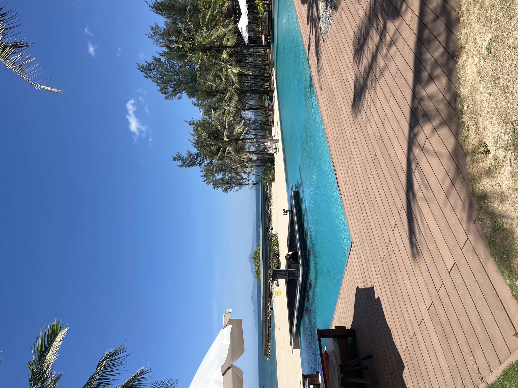 The image captures a beautiful and serene tropical resort scene in Đà Nẵng, Vietnam, under a bright, clear blue sky dotted with a few wispy white clouds. The perspective looks out towards the ocean from a poolside vantage point.
In the foreground, a large, rectangular infinity swimming pool with vibrant turquoise water runs parallel to a warm brown wooden deck. The deck, composed of long planks, shows distinct shadows cast by unseen objects, likely palm trees, indicating strong overhead sunlight. To the far right, a strip of dry, light-brown grass or earth marks the edge of the paved area.
Beyond the pool and deck, a pristine strip of light-colored sandy beach extends along the coastline. A dense line of tall, verdant trees, primarily palm trees and some conifer-like evergreens, separates the resort grounds from the sand, creating a natural screen.
In the mid-ground, the calm, blue expanse of the ocean stretches out to the horizon. Distant mountains are visible across the water, their peaks softened by a slight atmospheric haze. On the left side of the frame, the fronds of a palm tree are visible at the top and bottom, and a large white beach umbrella is partially seen in the lower-left corner, suggesting a seating area. Within the pool, a dark, built-in structure, possibly a sunken bar or seating area, adds to the luxurious feel.
The time of day appears to be sunny midday or early afternoon, given the clear skies and sharp shadows. The weather is perfect for a tropical vacation. The scene evokes a strong sense of relaxation, tranquility, and luxury, characteristic of a high-end coastal resort. There is no discernible text visible within the image.
