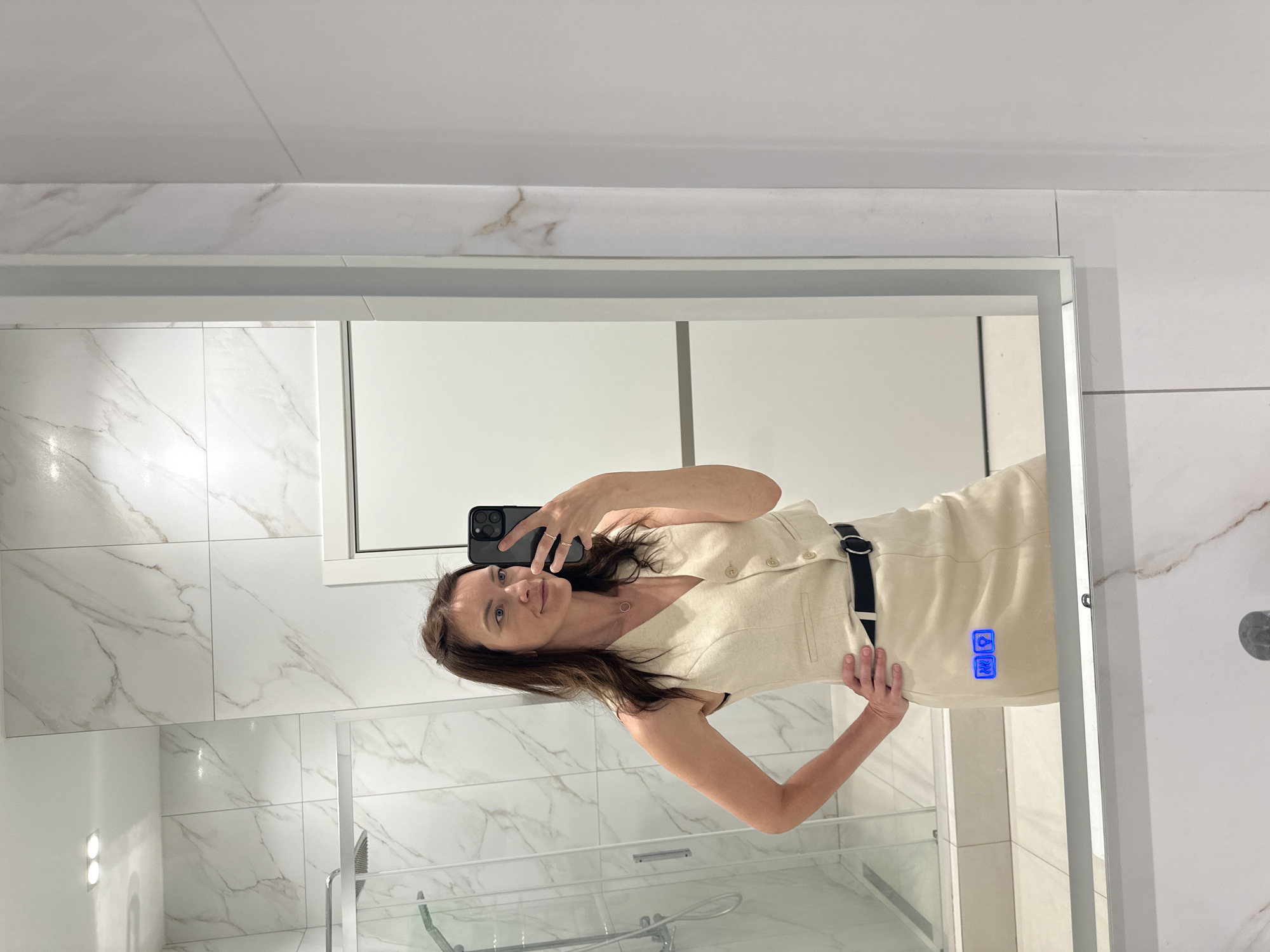 A woman is taking a mirror selfie in a bathroom. The bathroom has white marble-patterned tile walls, a glass shower enclosure, and a large mirror with a white frame. The woman is holding her smartphone up with one hand, and has the other hand on her hip. She is wearing a light-colored sleeveless dress with a black belt. The lighting in the bathroom is bright. The location is Przykamensky, Russia.