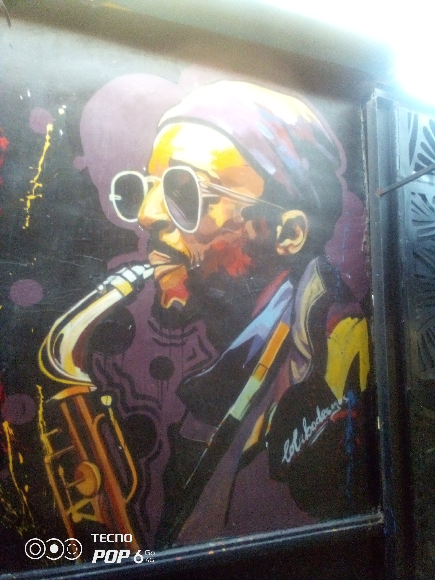 The image shows a painting of a man playing a saxophone. The painting is vibrant and uses bold colors, with the man's face rendered in yellow and orange tones, and his sunglasses in white. He is wearing a headband or cap and a jacket. The saxophone is depicted in golden yellow and orange hues. The background is dark with purple shapes and yellow splatters. There is a signature in white cursive script on the right side of the painting. A portion of a dark, ornate metal gate is visible on the far right edge of the image. A Tecno Pop 6 4G watermark is present in the bottom left corner. The painting appears to be on a wall or surface that has some texture. The lighting suggests it might be an indoor or shaded outdoor location. The image is blurry and has some glare. While the painting depicts a scene that might be found in a city like Howayi, Nigeria, the image itself does not provide any direct visual information to confirm its specific location.