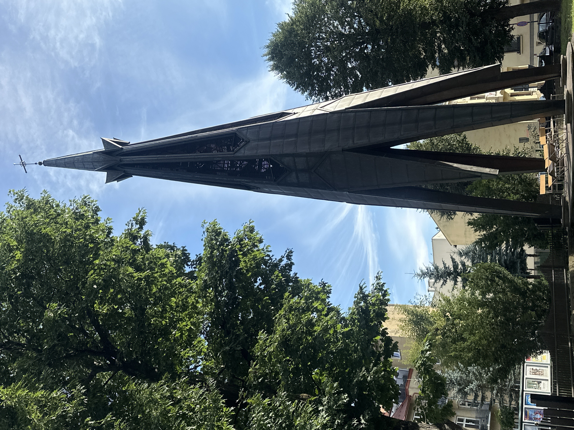 The image captures the Monument to the Fallen and Murdered in the East in Nowy Sącz, Poland. The monument is a tall, dark metal spire with an open, angular design, culminating in a cross at its apex. It is positioned in a public space, with trees on either side and behind it. A large, leafy tree dominates the left side of the frame, its branches extending towards the spire. In the background, several buildings, typical of a European city, are visible, some with apartment windows and balconies. The sky is bright blue with wispy white clouds, suggesting a clear day. In the foreground, at the base of the monument, a circular platform is visible, with several empty chairs and small tables arranged on it. The ground around the monument is covered in grass. The overall scene conveys a sense of solemnity and remembrance.