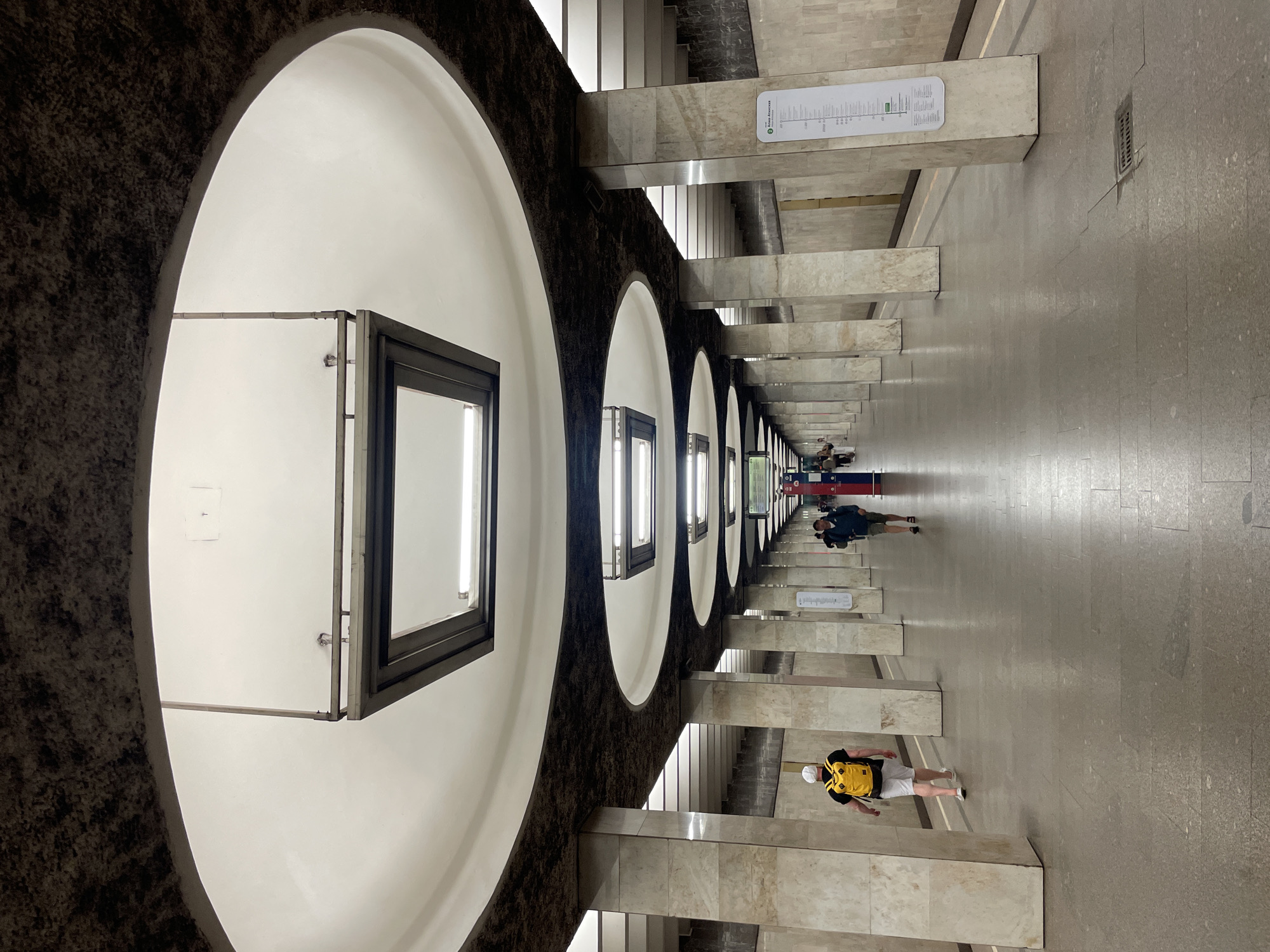 The image depicts an underground subway station in Moscow, Russia. A long corridor is lined with a series of large, oval-shaped arches. Within each arch hangs a square or rectangular light fixture. The walls of the station are clad in light-colored marble or tile. Several people are walking along the corridor. A man with a yellow backpack and white hat is walking in the direction of the camera. A couple or group of people are walking away from the camera. Informational signage is present on the walls. The floor is a light gray or beige color, and there are vertical support beams placed intermittently along the corridor.