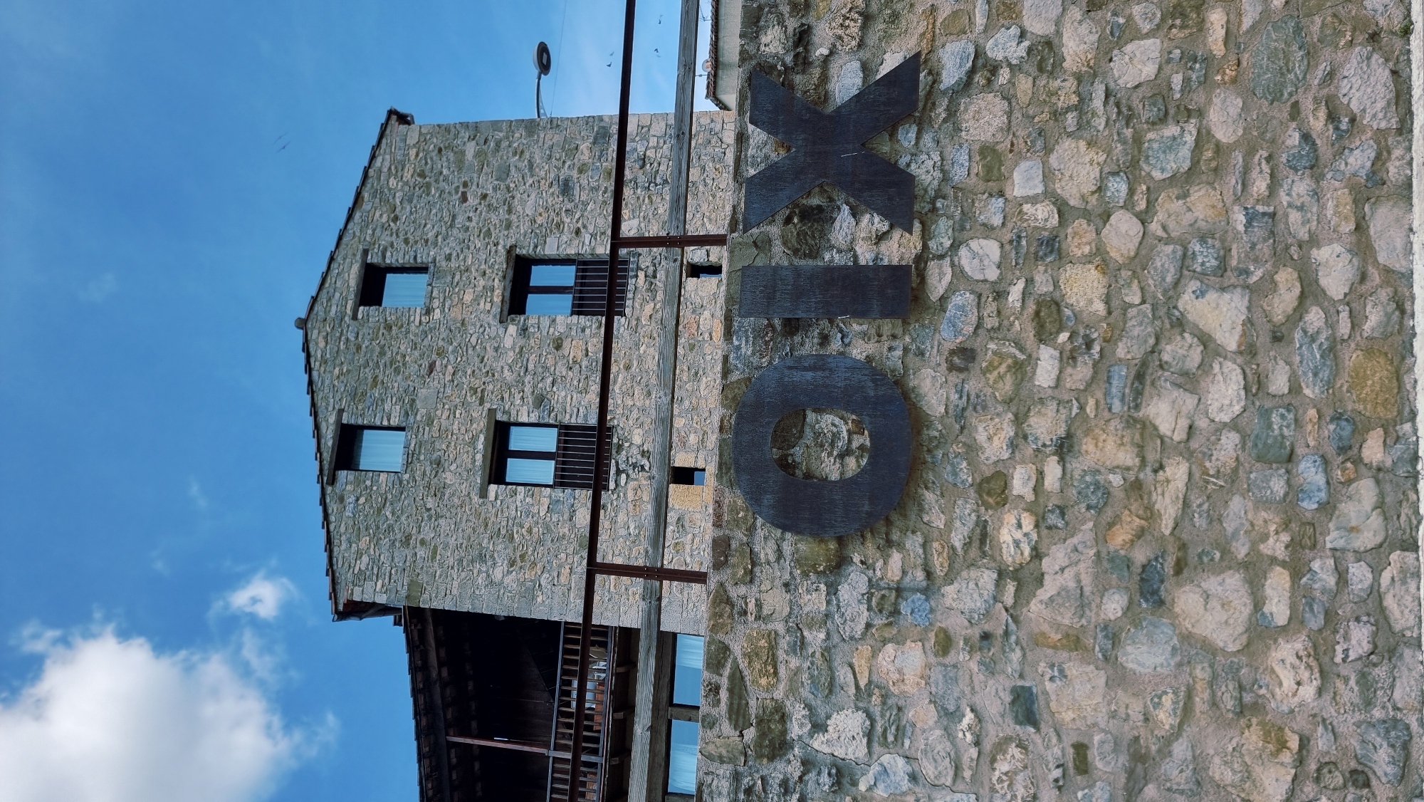 The image shows a building constructed with rough, grey stones. The building has a sloped roof and several square windows with dark frames. Some windows have metal grates. A dark wooden balcony or overhang is visible on the lower part of the building. To the right of the building, a large, dark metal sign is mounted on the stone wall. The sign consists of three separate letters: "X", "I", and "O", with the "X" being the largest and positioned above the other two. The letters appear to be made of thick, dark metal. The background is a bright blue sky with scattered white clouds, suggesting it is daytime. The image is oriented vertically. There are no people visible. The setting appears to be an outdoor architectural scene, possibly a historical building given the stone construction. The visible text on the sign spells out "XIO".