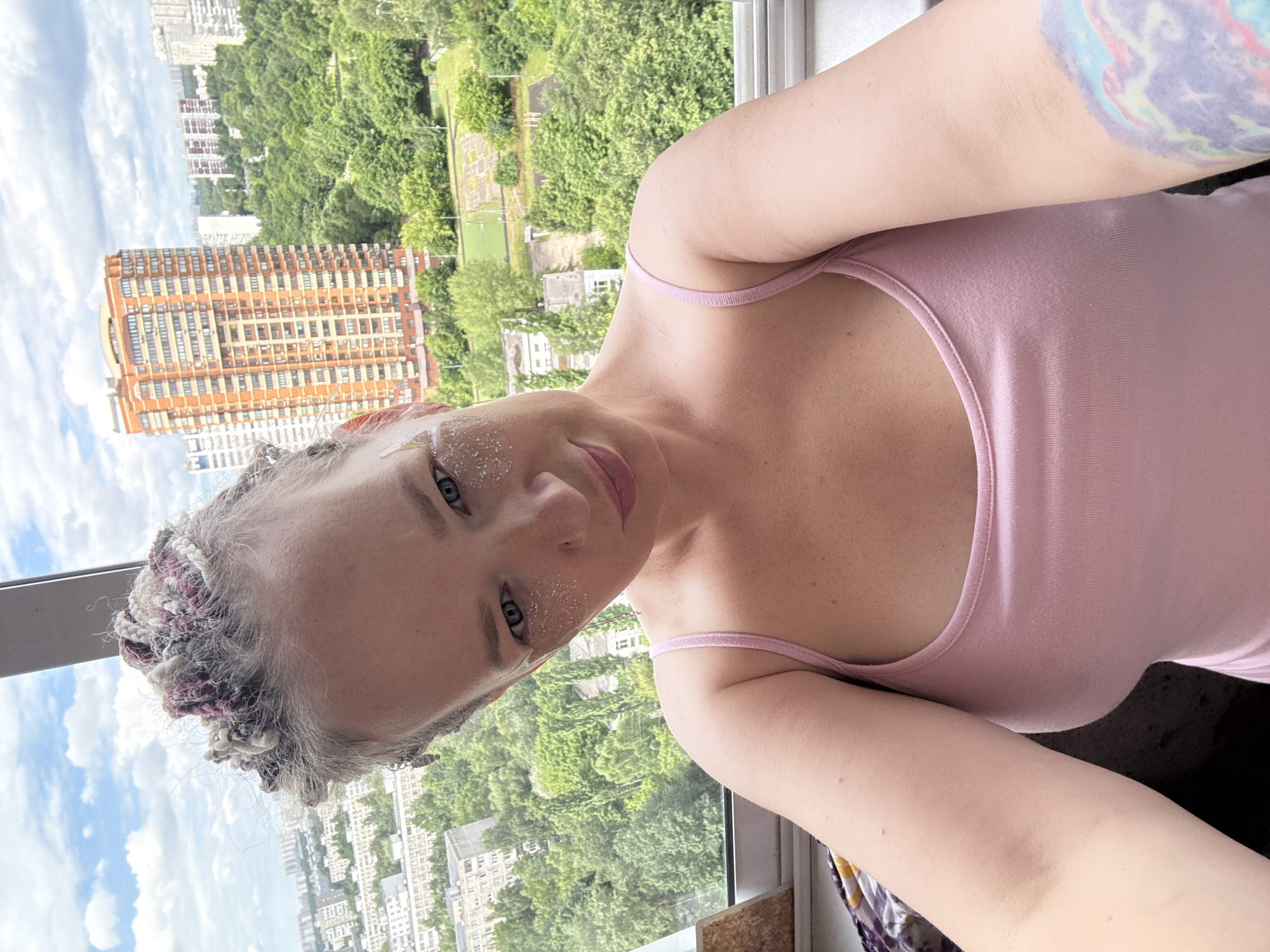 A young woman with braided hair and glitter on her face is taking a selfie indoors with a view of a city. The woman has fair skin and light eyes, and she is wearing a pink tank top. Her hair is styled in braids with streaks of pink and gray. She has glitter applied to her cheekbones and eyelids. Behind her, through a window, a city skyline is visible, with tall apartment buildings and lush green trees. The sky is cloudy and blue. The overall impression is of a casual, sunny day. The scene appears to be a residential area of Moscow, Russia, given the characteristic architecture of the buildings and the abundance of greenery.