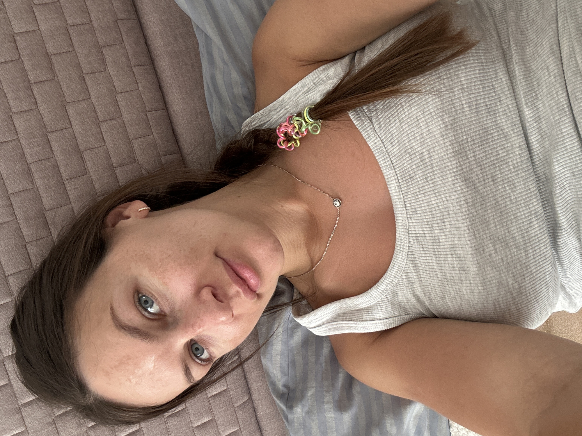 A woman with brown hair pulled back in a ponytail with colorful hair ties lies on a bed with a taupe textured headboard and a light blue striped duvet. She is wearing a light-colored tank top and a delicate necklace. The woman is looking directly at the camera with blue eyes and a slight smile. The scene appears to be indoors. There is no indication this photo was taken in Donji Pobori, Montenegro.
