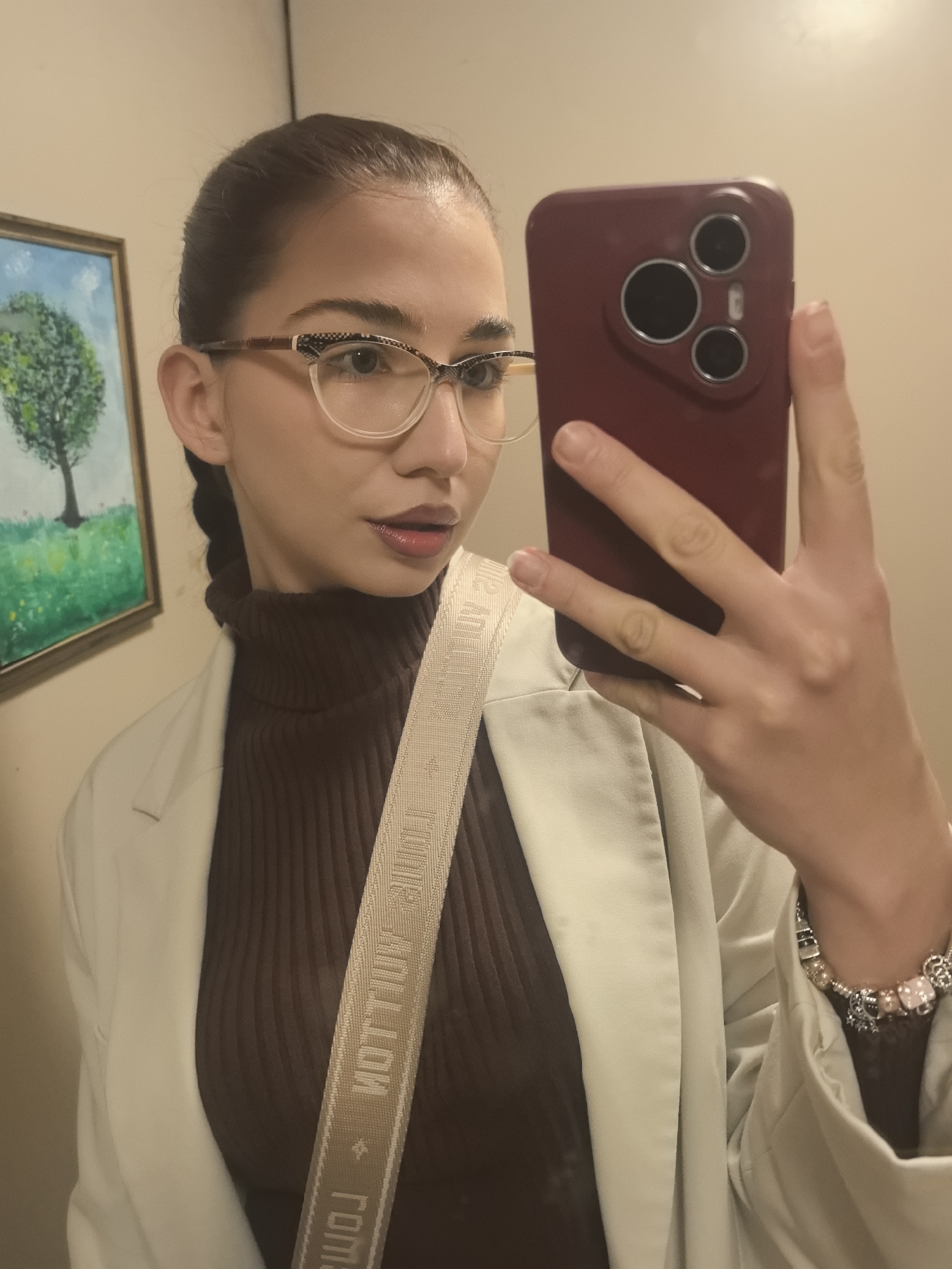 A woman stands indoors, taking a selfie with a red-cased smartphone. She has brown hair pulled back, wears glasses, a brown turtleneck sweater, and a light-colored blazer. A patterned strap crosses her body. She wears a charm bracelet on her left wrist. A framed picture hangs on the wall behind her. The location cannot be determined as Stara Zagora.