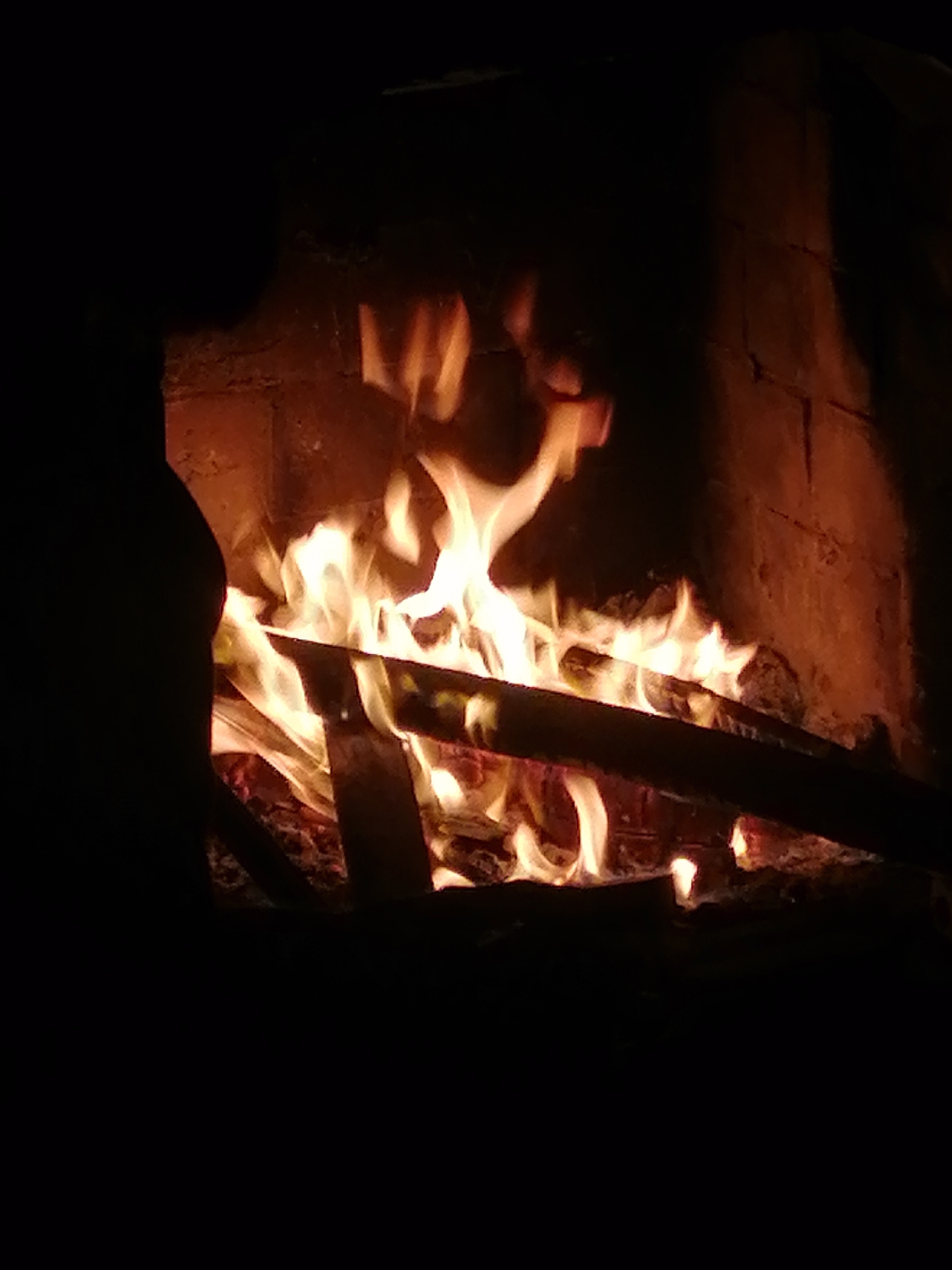 This image captures a warm and cozy scene of a crackling fire within a stone or brick fireplace, likely taken indoors during the evening or night. Bright, lively flames in shades of orange and yellow engulf several pieces of dark firewood, casting a warm, flickering light on the textured interior walls of the fireplace. Much of the surrounding area is immersed in deep shadow, highlighting the luminosity and heat of the fire. On the left side of the frame, a dark, indistinct silhouette, possibly a person or a structural part of the fireplace, is partially visible. No visible text is present. The overall atmosphere conveys comfort, tranquility, and warmth, suggesting a pleasant retreat from cold weather. This peaceful moment is set in Lạc Dương, Vietnam.