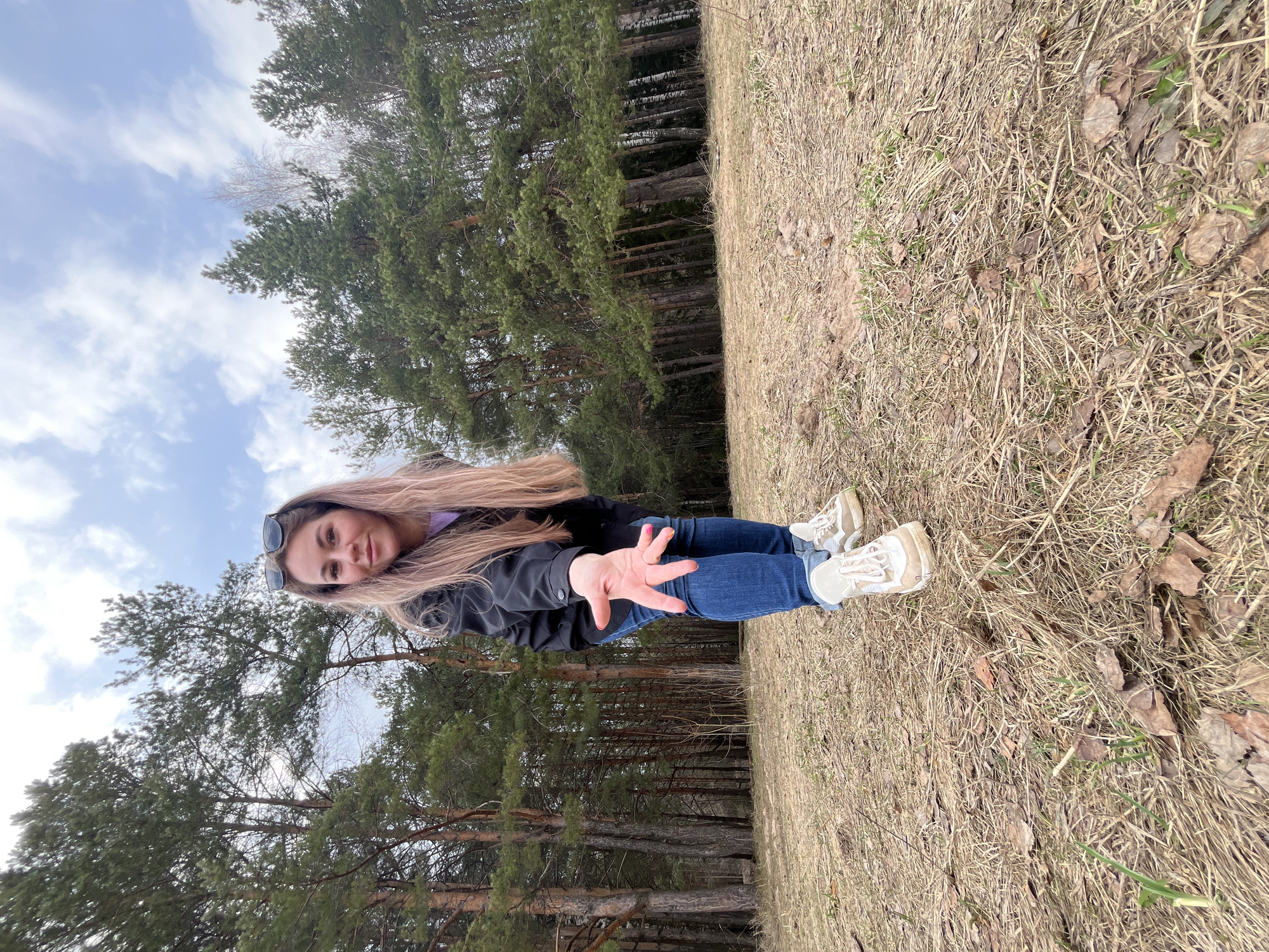 A young woman stands in a grassy clearing adjacent to a dense forest, likely within the city of Chaikovsky, Russia, given the prompt's instructions. The forest is characterized by tall, coniferous trees. The woman is wearing blue jeans, a dark jacket, and light-colored sneakers. Her long hair is light brown or blonde. She is looking directly at the camera, her right hand extended outwards. The sky is visible in the upper portion of the image, showing a partly cloudy pattern. The ground is covered with dry grass and some leaves, suggesting a transitional season, possibly spring or autumn.