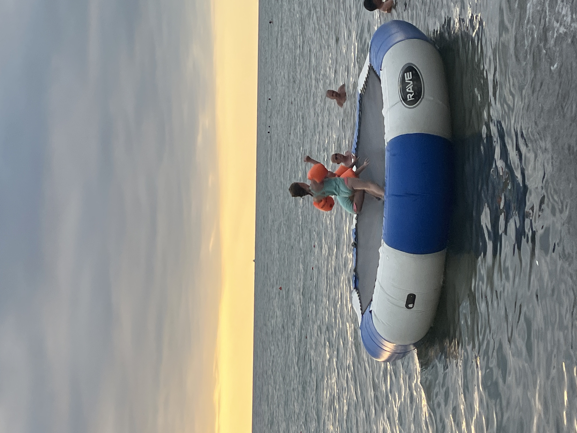 The image depicts a scene at Ureki, Georgia, a city known for its magnetic sand beaches. In the foreground, an inflatable trampoline floats on the water, with two young children, one wearing orange arm floaties and the other in an aqua swimsuit, actively playing on it. Several other individuals, appearing to be adults, are visible in the background, wading or swimming in the sea. The sky above is overcast, with a soft, warm glow of sunset visible on the horizon. The overall atmosphere suggests a recreational and relaxed setting by the Black Sea.