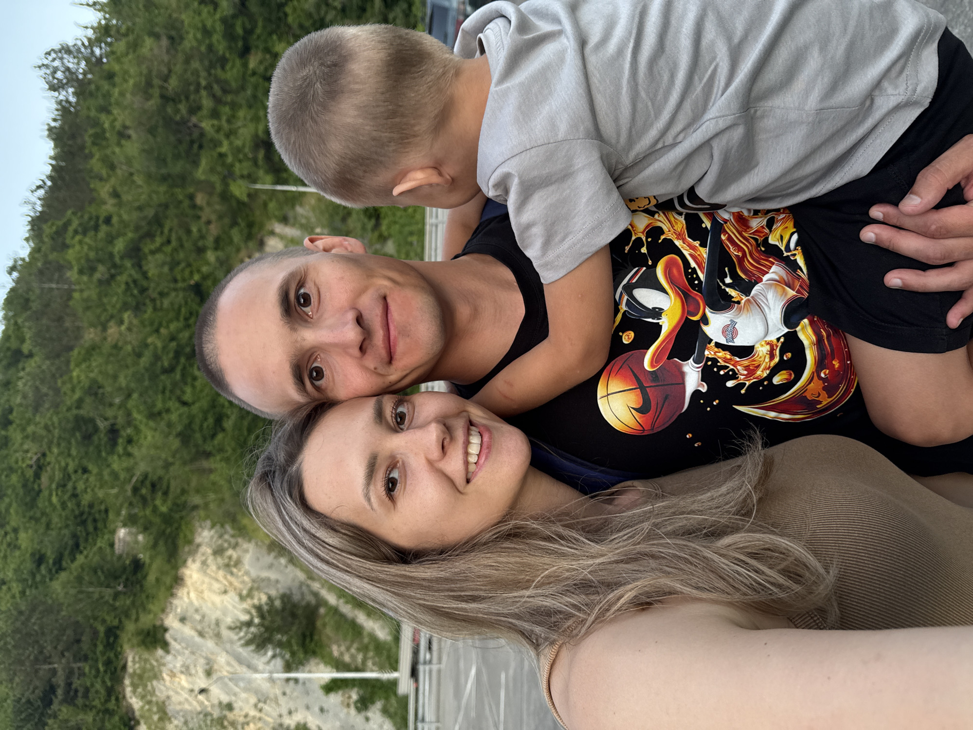 A medium shot captures a family of three, a man, a woman, and a young boy, posing for a selfie in what appears to be an outdoor, elevated setting overlooking a lush, tree-covered hillside. The man, with a shaved head and a slight smile, is in the center, holding the boy who is turned away from the camera, embracing the man. The woman, to the left, smiles warmly, her blonde hair framing her face. She has her arm around the man. The man wears a black t-shirt with a cartoon graphic of Daffy Duck playing basketball. The boy is in a light grey t-shirt, and the woman wears a light brown ribbed tank top. The background reveals a steep, rocky slope dotted with trees and vegetation, suggesting a natural or semi-natural environment, possibly a park or a scenic overlook. A road and some railings are visible in the mid-ground. The lighting indicates it is likely daytime, with a soft, natural light illuminating the scene. The location is identified as Nebug, Russia.