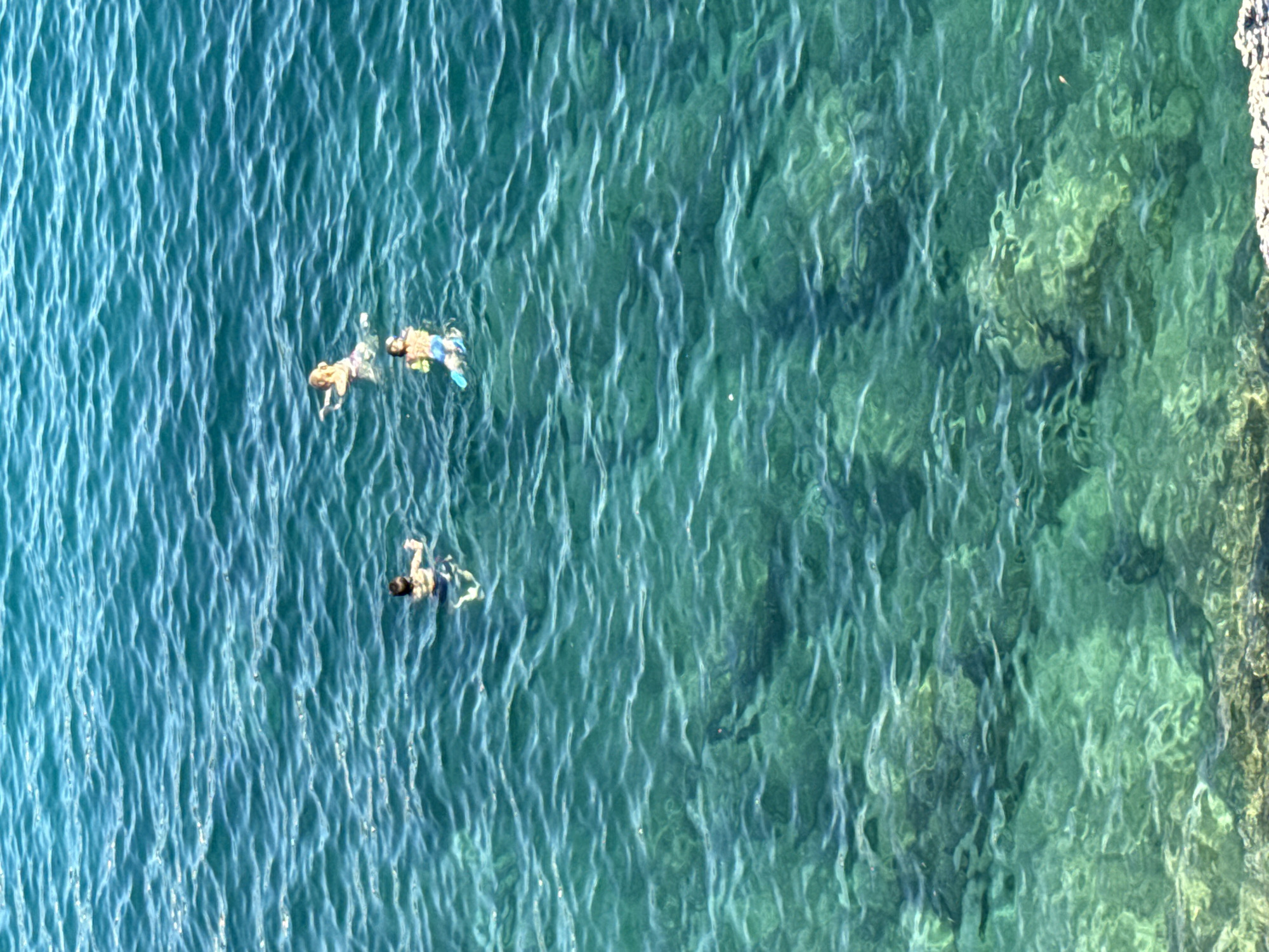 The image shows a group of people swimming in the clear turquoise waters of the Adriatic Sea near Kamenovo, Montenegro. The sun is shining, creating a shimmering effect on the water's surface. In the foreground, rocks are visible beneath the water, indicating the shallow depth of the area. The people are engaged in leisurely swimming and snorkeling, enjoying the serene beach environment.