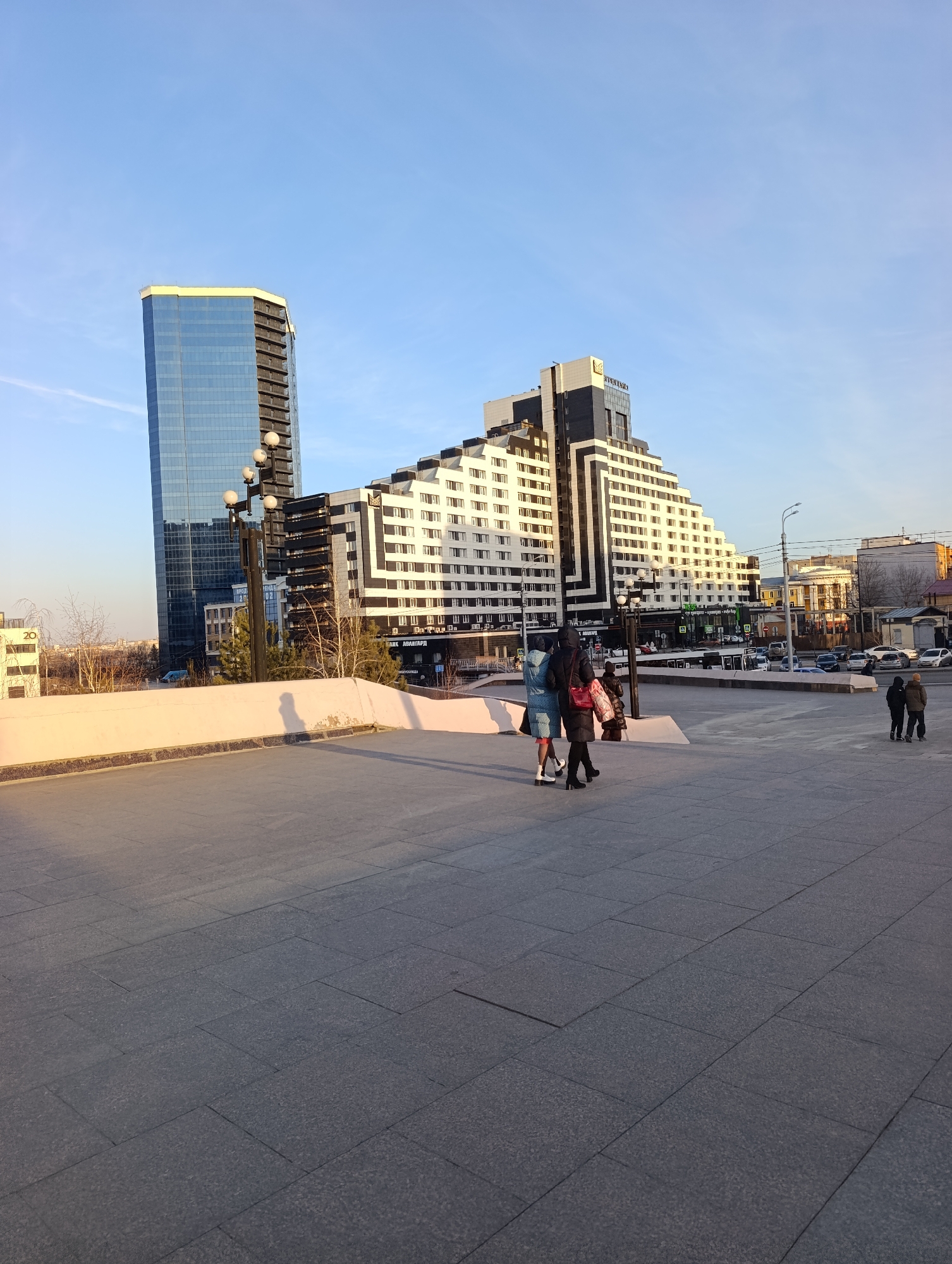 The image depicts a city scene in Krasnoyarsk, Russia. The prominent architectural features include two tall buildings: one a modern glass skyscraper, the other a pyramid-shaped hotel or office building with alternating horizontal stripes of white and dark colors. Several people are visible in the foreground on a stone-paved plaza. A group of three people walks away from the camera, and another group of two individuals stands in the distance. Cars and other buildings are visible in the background across a street, indicating an urban environment. The time of day seems to be late afternoon or early evening, judging by the long shadows cast on the plaza.