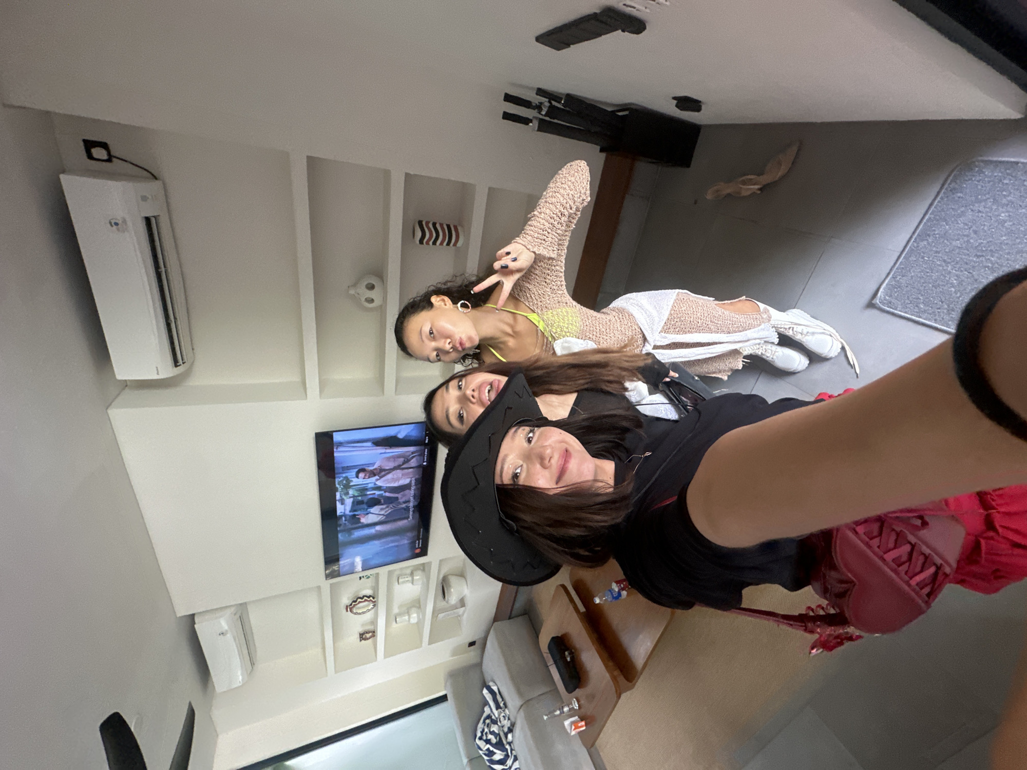 Three women are posing for a selfie in an indoor setting. The woman in the foreground is wearing a black shirt, a black cowboy hat, and carrying a red purse. The second woman is partially visible behind her. The third woman is in the background, wearing a yellow top and a beige knitted cover-up, making a peace sign. The location appears to be a living room with a TV mounted on a wall with built-in shelves containing decorative items. An air conditioning unit is also visible on the wall. There is a couch in the room, and the flooring appears to be tile. Based on the objects and architectural style, the location is likely in North Kuta, Indonesia.