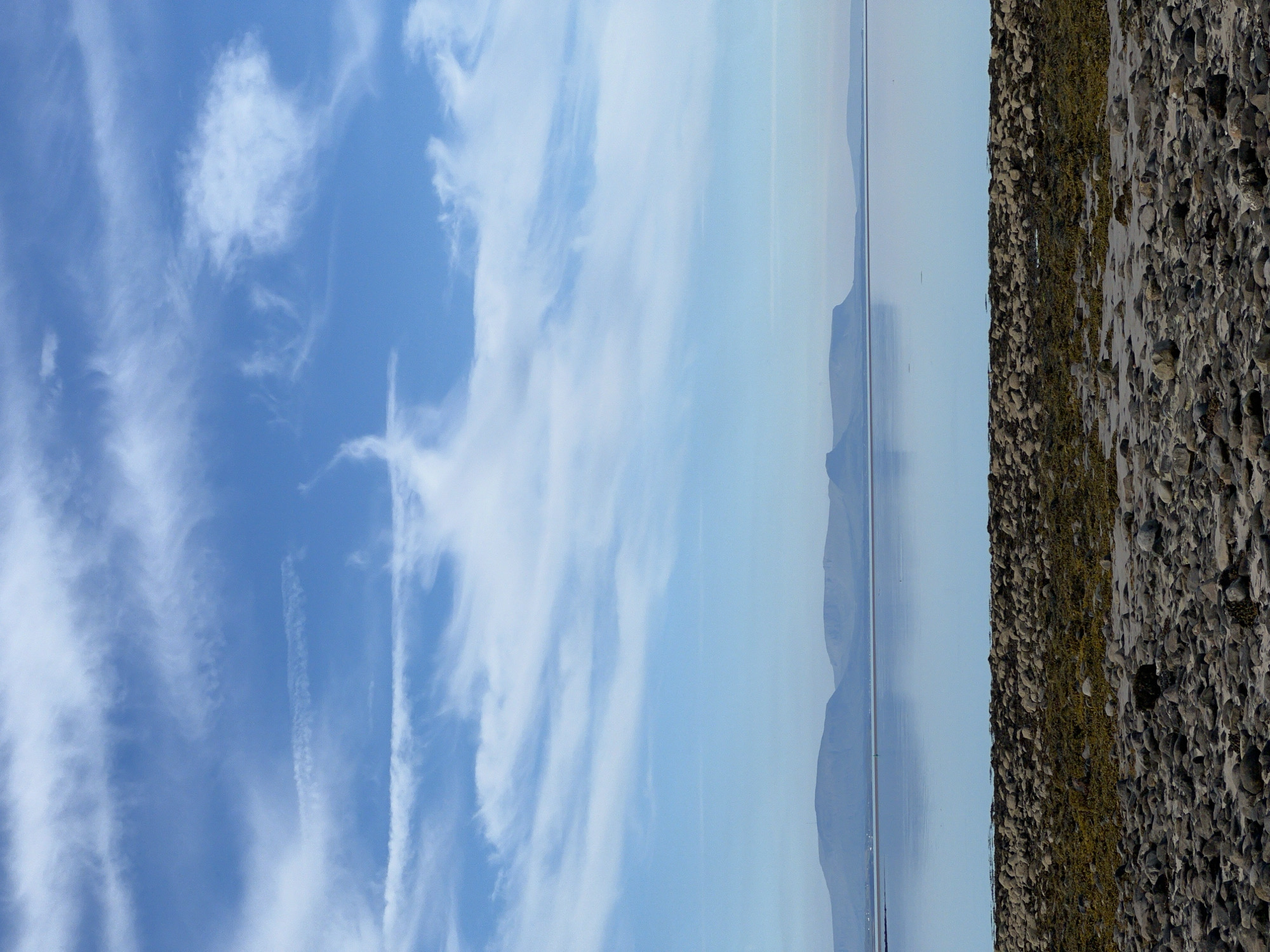 This media file, taken in Mountcharles, Ireland, captures a serene natural landscape. The image is oriented vertically, with the sky occupying the left side and the ground/sea on the right. Thin, wispy white clouds streak across a bright blue sky, suggesting a clear day with some atmospheric movement.
The right side of the image transitions from a calm, reflective body of water to a rocky shoreline. The water appears still, mirroring the soft blue of the sky and distant, hazy mountains. These mountains form a gentle, undulating silhouette against the horizon, indicating a coastal or lakeside setting. The shoreline is covered in numerous grey and brown stones, interspersed with patches of green moss or seaweed, suggesting a rugged, natural beach.
There is no discernible activity or event taking place; the scene is static and peaceful. The lighting suggests it is daytime, likely late morning or early afternoon given the brightness. The weather appears fair and calm. The overall mood conveyed is one of tranquility and natural beauty, characteristic of a quiet, scenic location. No text is visible in the image.