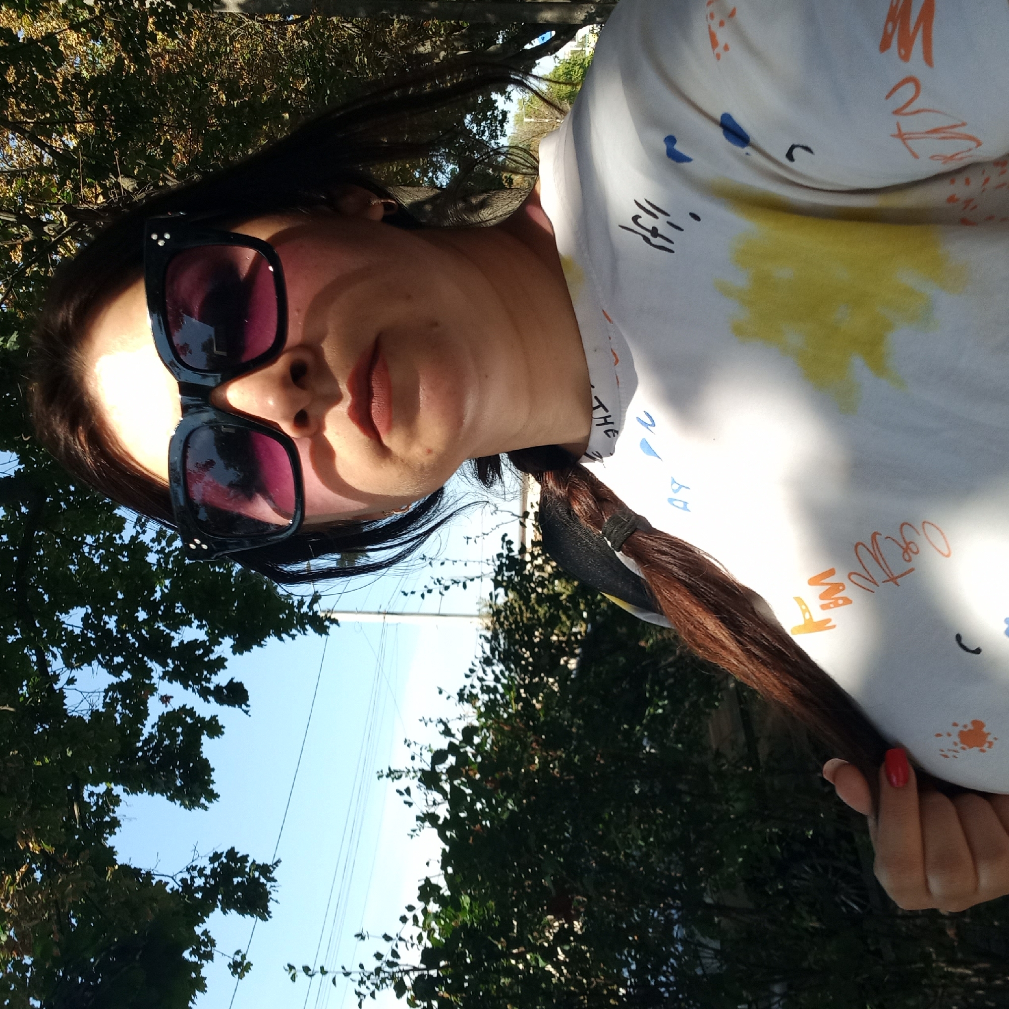 This is a selfie taken outdoors, likely in a park or along a street lined with trees, as indicated by the foliage and dappled sunlight. The subject is a woman with dark hair, wearing large, square-shaped sunglasses with a black frame and purple lenses. She has on a white t-shirt with a colorful abstract design featuring splatters of yellow and orange, as well as some black and blue markings that appear to be text or symbols. Her hair is pulled back into a braid that hangs over her shoulder, and she is touching it with her left hand, which has red nail polish. The background consists of green trees, blue sky, and power lines, suggesting an urban or suburban setting. The lighting indicates it is daytime. The specific location in Kropyvnytskyi, Ukraine, cannot be determined from the visual information alone.