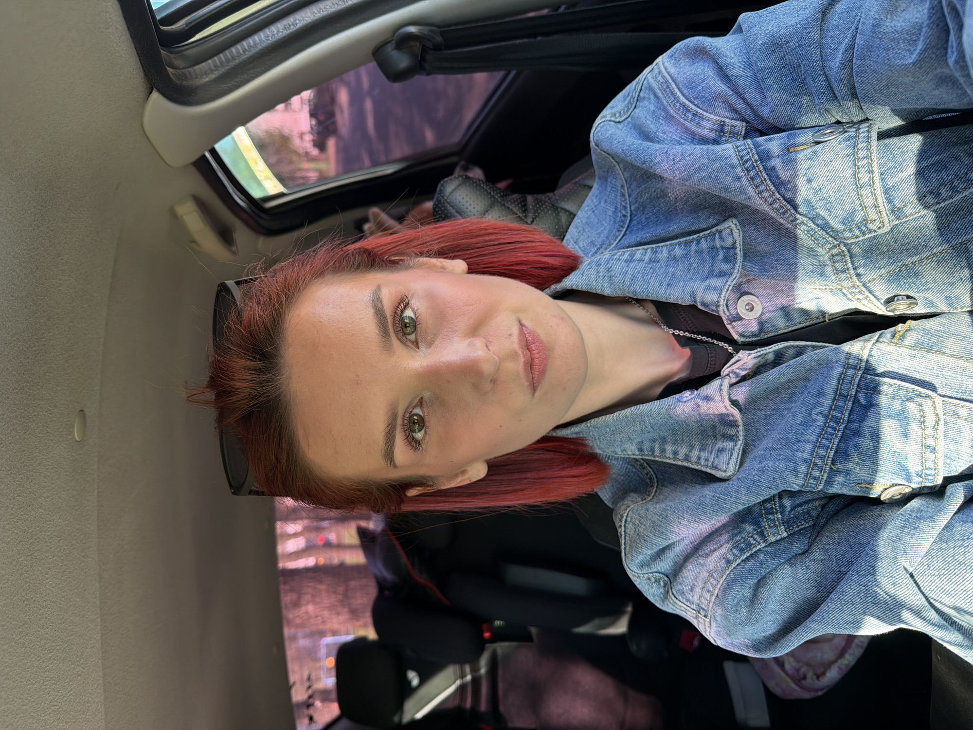 A woman with red hair is sitting in a car. She is wearing a denim jacket and a necklace. The car has a sunroof. The city of Chaikovsky, Russia is visible through the sunroof.