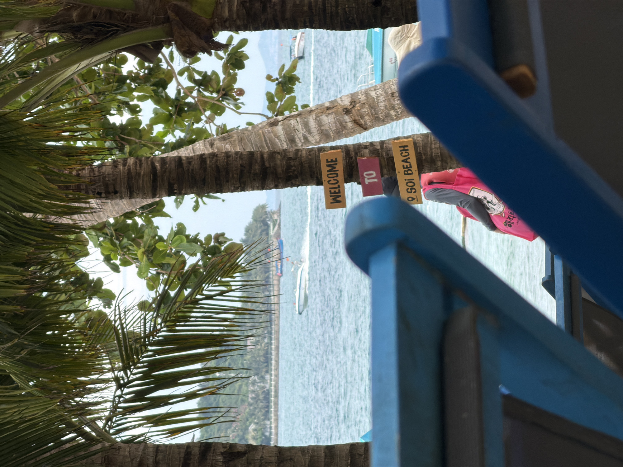 This image captures a vibrant tropical beach scene, likely in Nam Nha Trang Ward, Vietnam. The perspective places the viewer from behind a series of large palm trees, whose rough, segmented trunks and lush green fronds dominate the foreground and frame the view of the sea.
Beyond the palm trees, a calm, light blue-green ocean extends across the midground. Several small boats are visible on the water, including a distant motorboat and another closer watercraft, hinting at recreational activities. A faint landmass or coastline is visible on the far horizon beneath a bright, clear sky.
A notable detail is a set of three small, rectangular wooden signs hanging from one of the palm tree trunks. They read, from left to right, "WELCOME", "TO", and "SOI BEACH" (with a location pin icon), clearly indicating the specific beach location.
On the far right, parts of a blue structure, possibly a beach chair or outdoor furniture, are visible and slightly out of focus, framing the scene. Behind these blue elements and near the "SOI BEACH" sign, a bright pink item, which could be a bag or part of an outfit, is visible. It features some Korean text, including "해저" (hae-jeo), meaning "underwater" or "seabed".
The scene is bathed in bright daylight under sunny, clear weather conditions, suggesting a leisurely day at the beach. The overall impression is one of a relaxing tropical vacation spot.