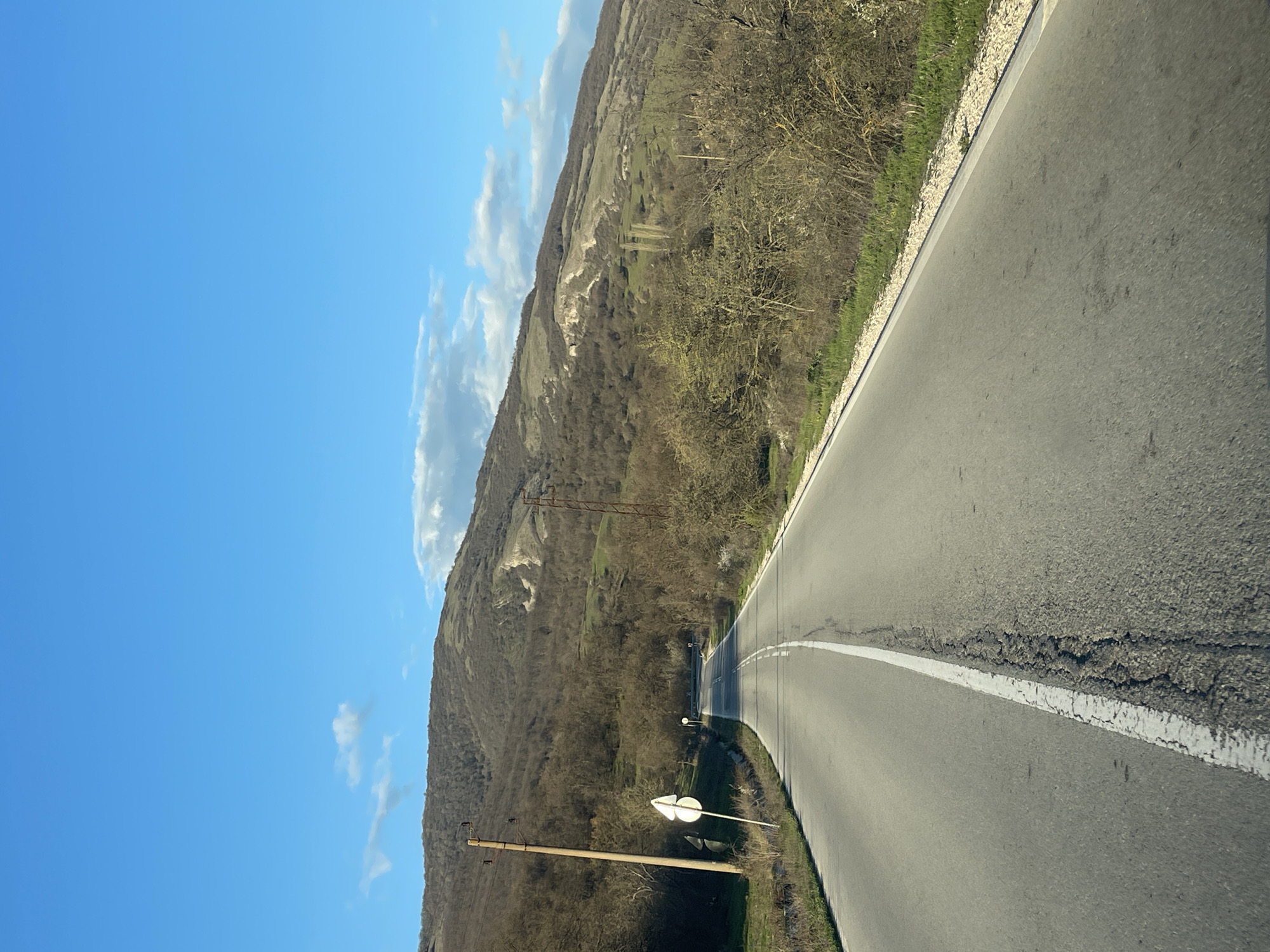The image shows an upward-sloping asphalt road with a central white dividing line, likely located in a rural or mountainous area, possibly near Kamchiya, Bulgaria. The road is adjacent to a wooded hillside with sparse vegetation and some exposed rocky areas. Overhead power lines are visible along the side of the road. The sky is blue with scattered white clouds. A road sign is visible on the left side of the road. The image appears to be taken from a moving vehicle.