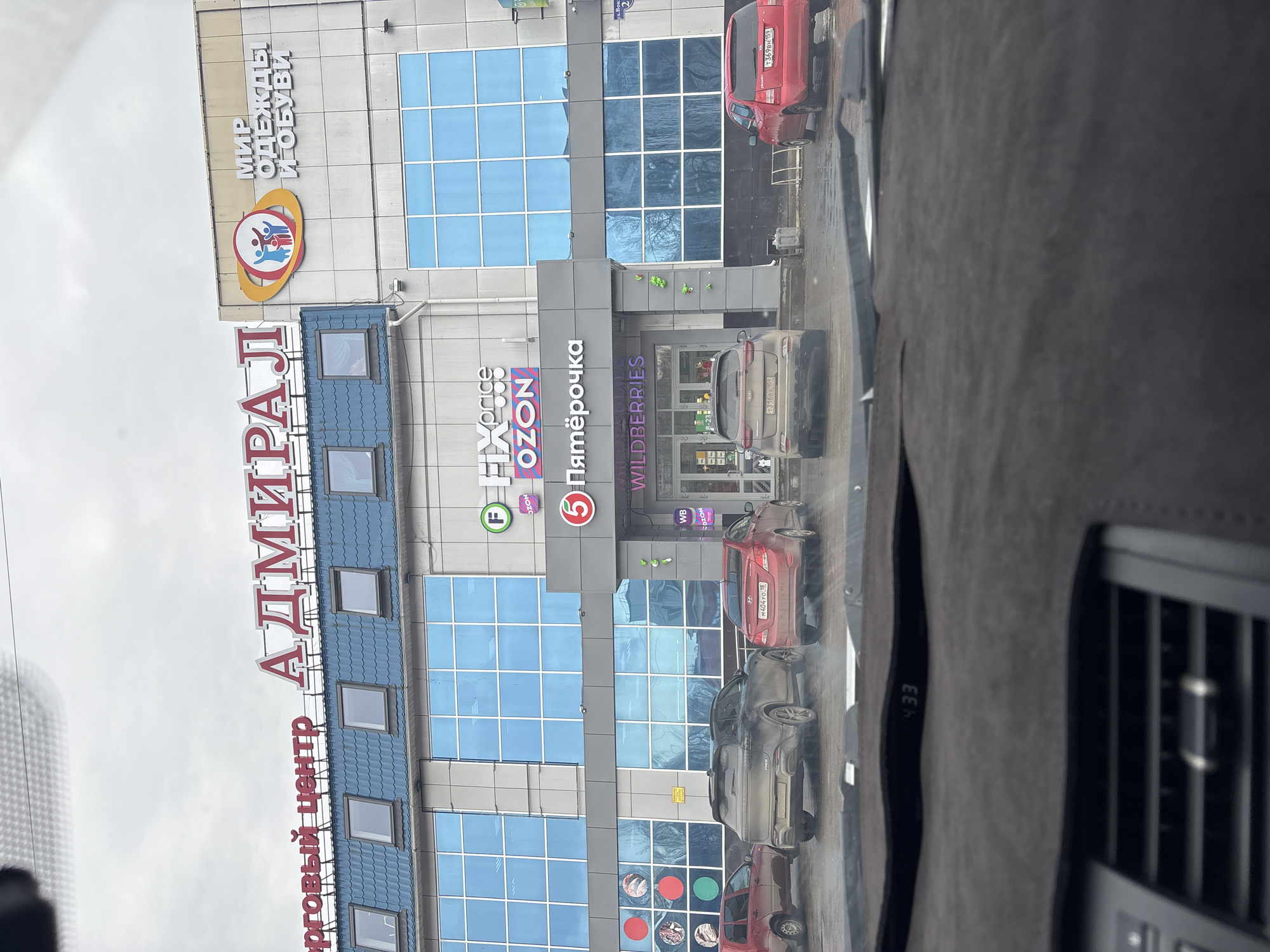 The image shows the "Admiral" shopping center in the city of Chaikovsky, Russia. The building has multiple tenants, including "Pyaterochka" (a grocery store chain), "Fix Price," "Ozon" (an e-commerce company), and "Wildberries" (another e-commerce company). A sign above reads, "Mir Odezhdy I Obuvi," translating to "World of Clothes and Shoes." Several cars are parked in front of the building. The photo was taken from inside a vehicle with the dashboard partially visible. The building is multi-story and predominantly blue and gray, with large window panels.