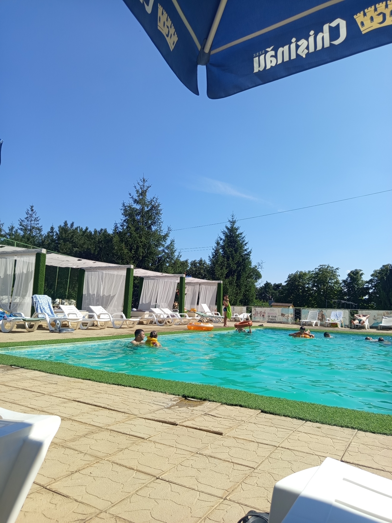 A bright, sunny day at a resort pool in Molovata Nouă, Moldova. In the foreground, a white plastic lounge chair is partially visible. The pool itself is a vibrant turquoise, with several people swimming and relaxing. Two individuals are in the water near the left side of the pool; one adult male appears to be holding a young child wearing a yellow flotation device. Other swimmers are dispersed throughout the pool, some with inflatable rings. The poolside is lined with white lounge chairs and cabanas draped with white curtains, offering shade. Artificial turf lines the edge of the pool. In the background, lush green trees and a clear blue sky dominate the horizon. A portion of a blue umbrella with white lettering is visible at the top of the frame.
