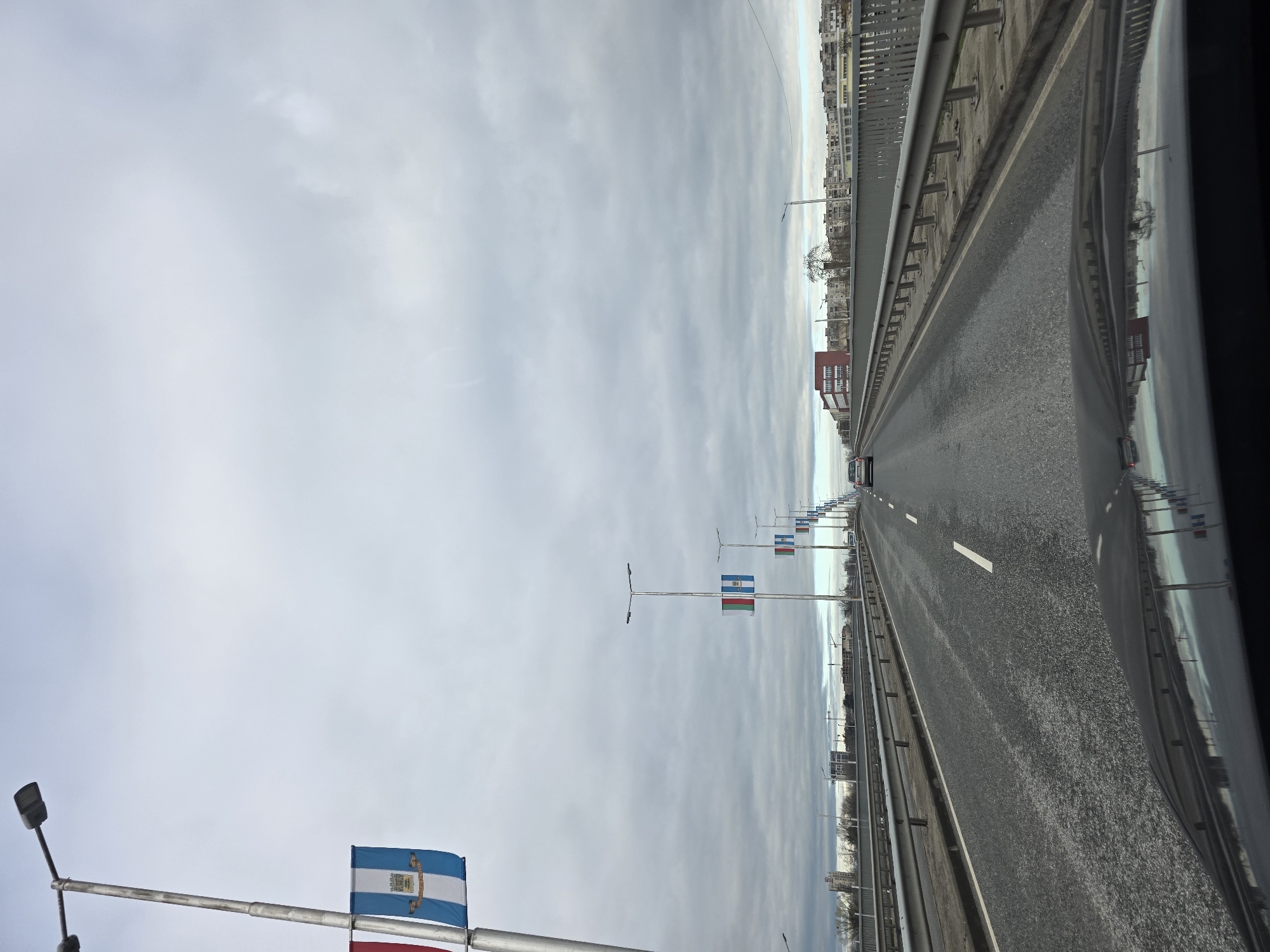 The image shows a section of the Maritsa Bridge in Plovdiv, Bulgaria. The view is from a vehicle traveling on the bridge. Several flagpoles with Bulgarian and Plovdiv flags line the road. The bridge spans the Maritsa River, and buildings are visible in the distance. The sky is overcast.