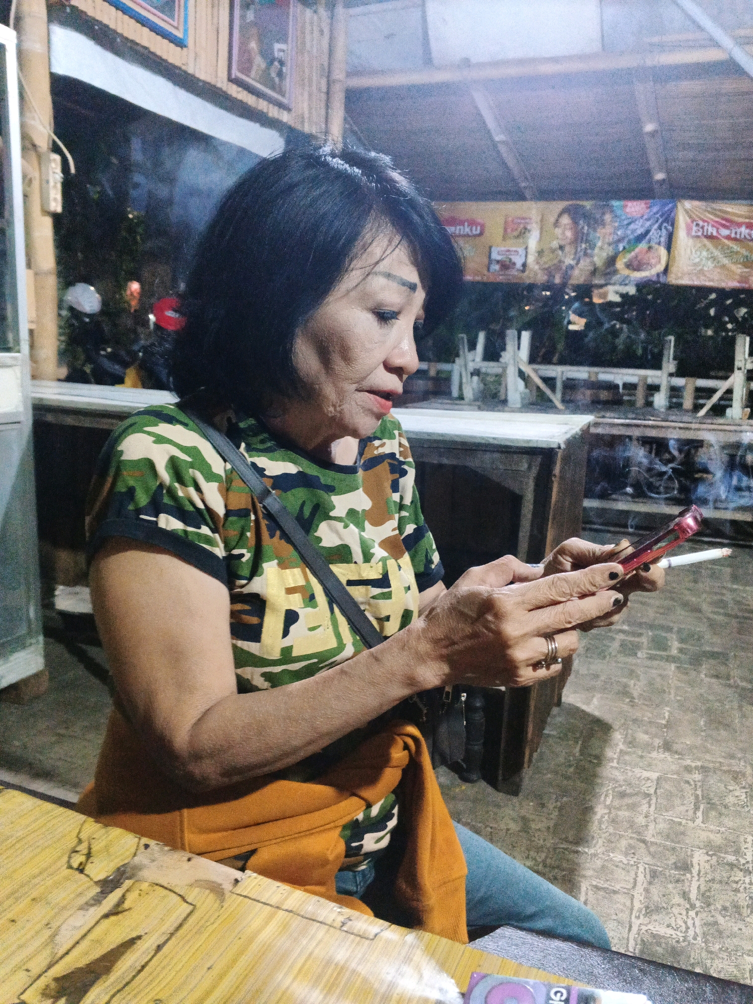 In Ungaran, Indonesia, a woman with short black hair and fair skin is seated outdoors at a table, holding a mobile phone and a cigarette. She is wearing a camouflage-patterned shirt and an orange sweater tied around her waist, with blue jeans. The woman is looking down at her phone, with the lit cigarette held between her index and middle fingers. The environment appears to be a casual, outdoor seating area with a rustic, wooden roof structure and bamboo accents. In the background, there are banners and what appears to be a fence or railing. The lighting suggests it is either dusk or nighttime.