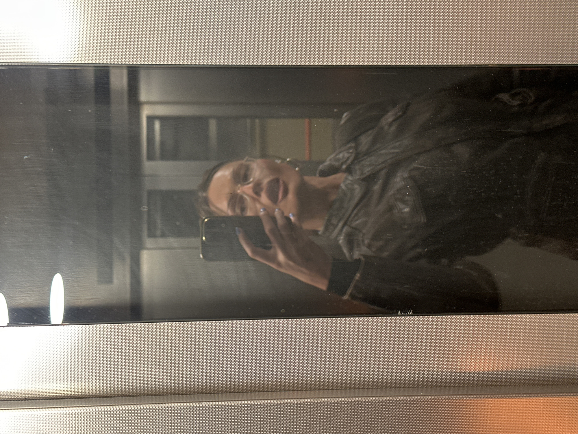 The image depicts a reflection in a glossy surface, possibly glass or a screen. The reflection shows a woman holding a phone, presumably taking a selfie. She is wearing a dark-colored jacket and has an open mouth, possibly talking or singing. The background in the reflection is indistinct but suggests an indoor environment, possibly an elevator. The surface reflecting the image has a textured border, possibly a microwave or other appliance. There is no information present in the image that would indicate the location in the city of London, United Kingdom.