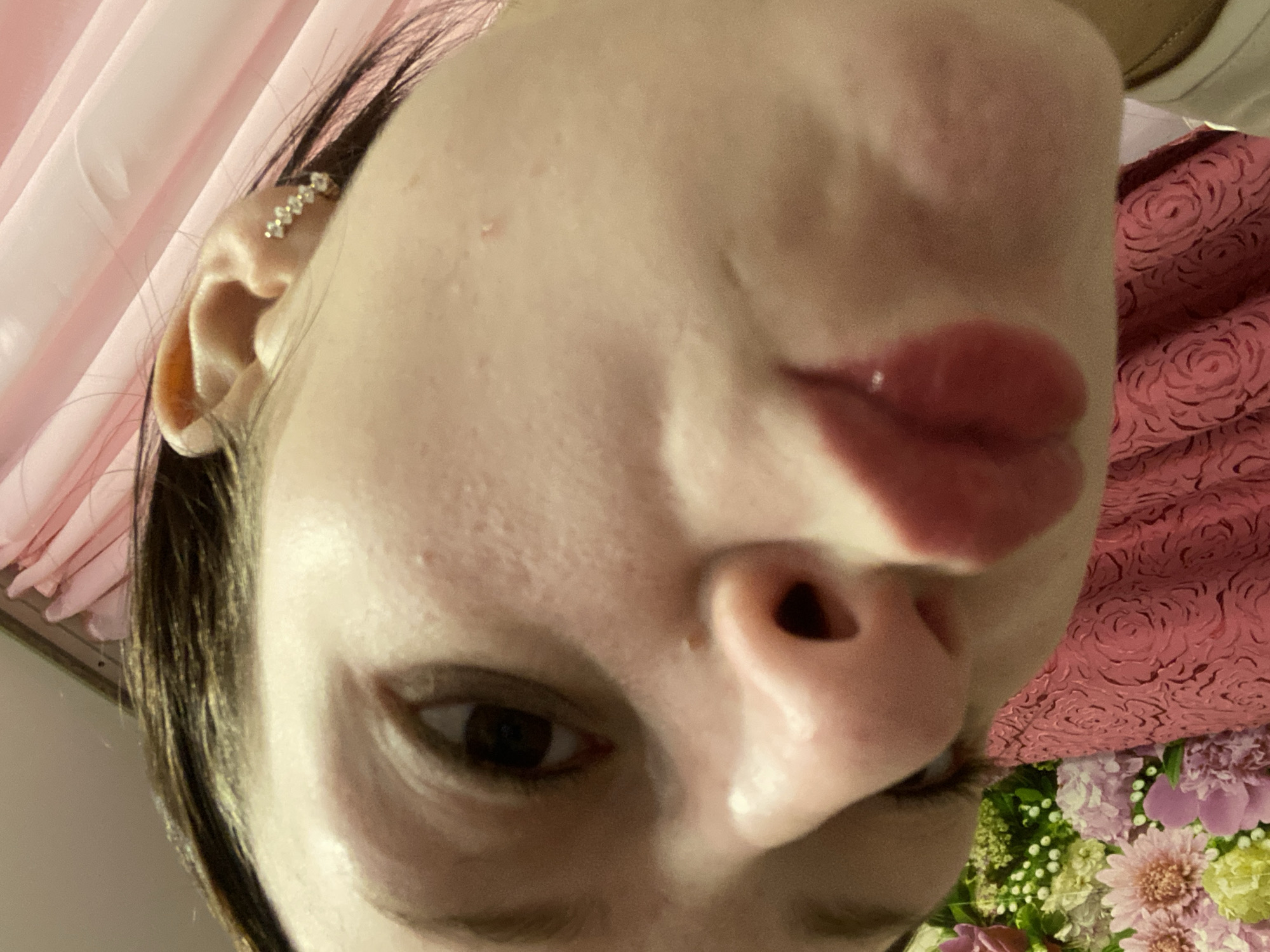 The image is a close-up, low-angle, portrait-style photograph of a young Caucasian female's face, tilted and partially obscured. Her dark hair is visible framing her forehead and left ear, which is adorned with a delicate, diamond-studded earring. Her eye is a prominent feature, looking directly at the camera. The lighting suggests an indoor setting, possibly a bedroom or dressing area, with a pink, draped fabric visible in the background and on her chest. A portion of a floral arrangement, featuring pink and yellow flowers, is visible in the lower left corner. The subject's lips are pursed, suggesting a playful or suggestive pose. The image does not contain any specific landmarks or indicators that would allow for its localization to the city of Медведево, Russia.