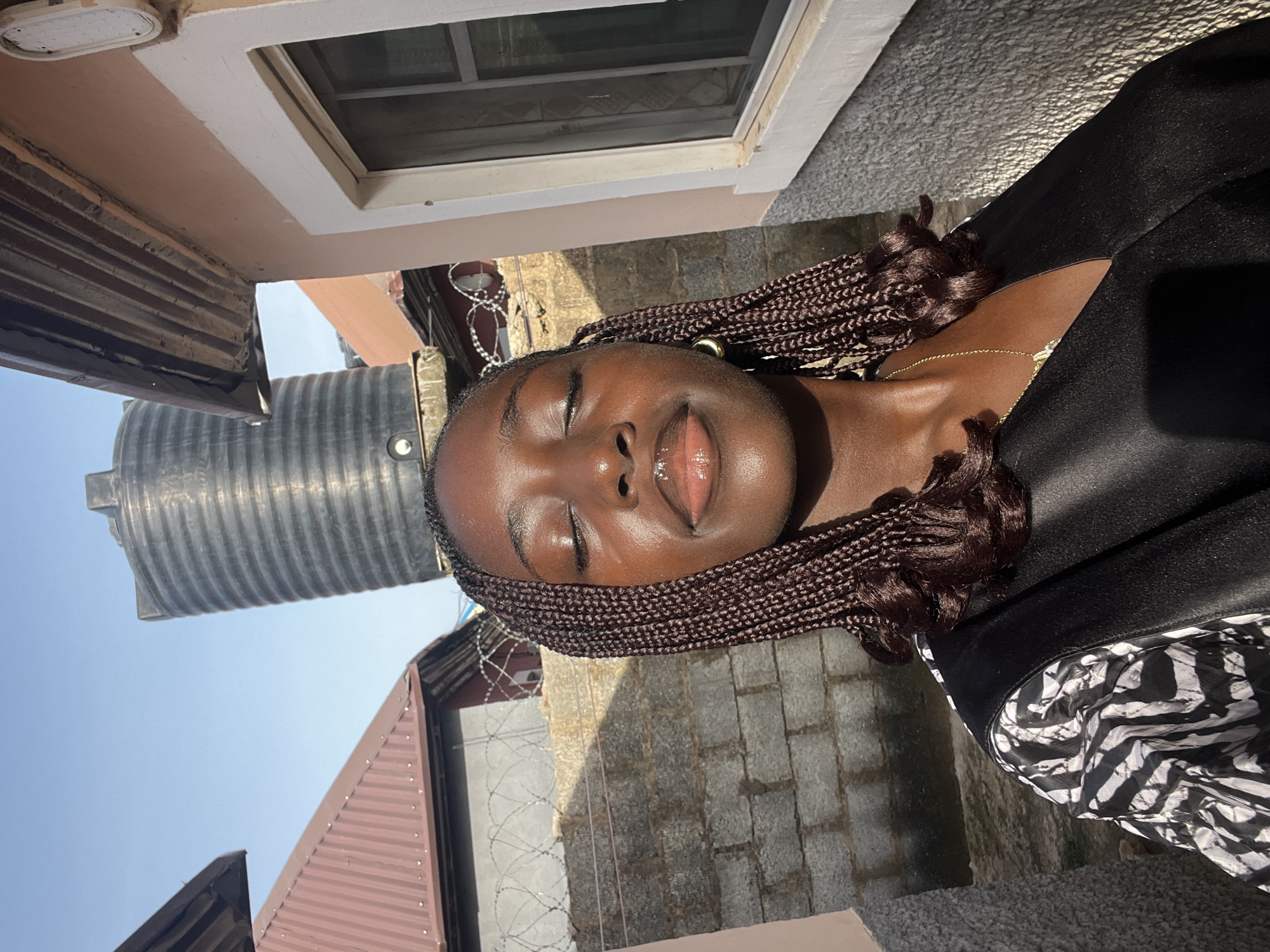 The image shows a young woman with dark skin and braided hair. Her eyes are closed, and her lips are slightly parted. She is wearing a black top with a zebra-striped pattern visible on the side. A gold necklace is visible around her neck.
The background includes buildings with brick and metal structures. A water tank is also visible.
While the image provides clues about the setting, it does not contain sufficient information to definitively confirm the location in Abuja, Nigeria. More specific details about the architecture or landmarks would be necessary for an accurate determination.