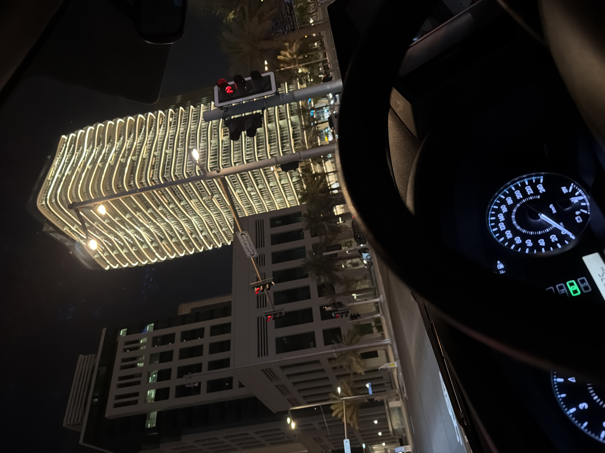 The image shows a view from inside a car at night. The car is stopped at a traffic light, indicated by the red light visible on the traffic signal in the foreground. Through the windshield, two modern buildings are visible. The building on the right is a tall, curved skyscraper illuminated by many horizontal lines of warm light. The building on the left is a darker, more angular skyscraper with visible windows. Palm trees line the street in front of the buildings. The car's dashboard is partially visible, showing a speedometer and other indicators. The overall scene depicts an urban environment at night, likely in a city with modern architecture.