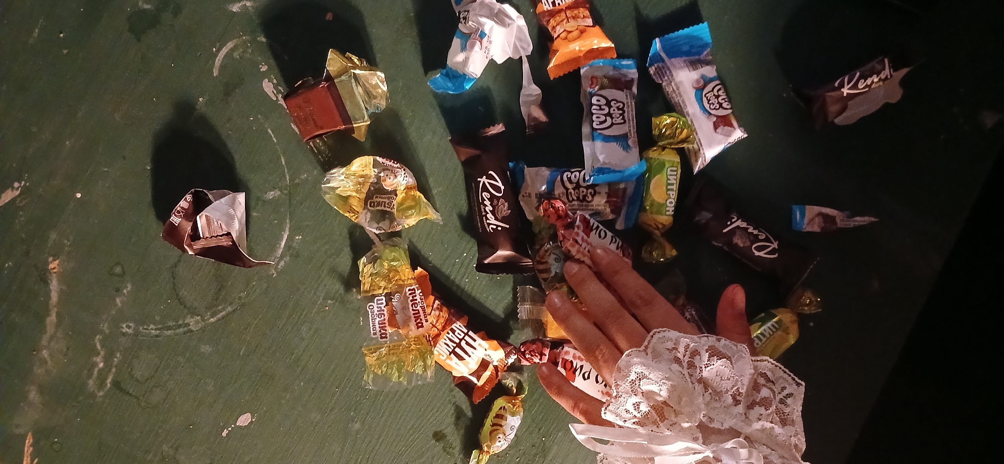 An overhead shot displays a collection of various candy wrappers and chocolates scattered on a dark green, worn surface, likely a table. A fair-skinned human hand, adorned with a white lace cuff, extends from the lower right towards the center of the frame, interacting with the pile of sweets.
Many of the wrappers are crinkled and partially open, indicating that candies have been consumed or are in the process of being unwrapped. The lighting suggests an indoor setting, possibly under artificial light, casting soft shadows. The green surface exhibits scuff marks and some circular stains, hinting at frequent use.
Visible text on the candy wrappers includes:
* "Kendi" and "Rendi" (on dark chocolate-like bars)
* "COCO ROPS" (on blue and white wrappers)
* "ЦИТРОН" (Citron, on a yellow and green wrapper)
* "СНiО РИС" (Snow Rice, on a clear wrapper with pink text)
* "НУГА АРАХИС" (Nougat Peanut, on an orange wrapper)
* "Озорная пчёлка" (Naughty Bee, on gold-colored wrappers)
The presence of these specific candy brands is consistent with the provided location context of Moscow, Russia. The scene captures a moment of casual indulgence in sweets.