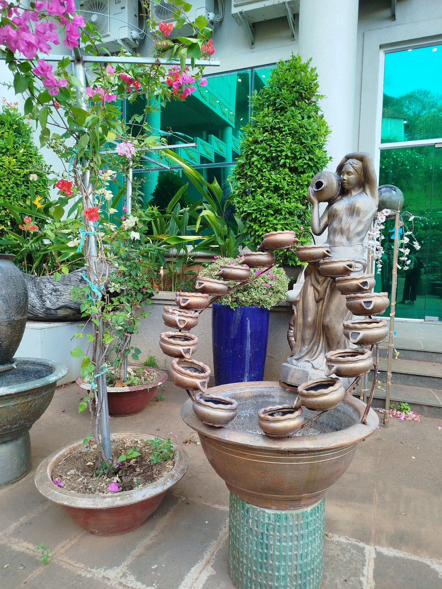 The image shows a garden scene in what is possibly Ho Chi Minh City, Vietnam. A multi-tiered fountain made of bronze-colored bowls dominates the view. It is positioned in front of a statue of a woman holding a jug. The fountain is tiered with smaller bowls cascading into a larger basin below, which is supported by a green-patterned stand. To the fountain's left are various plants in pots, including one with pink bougainvillea flowers climbing a metal trellis and another featuring green foliage. Behind the fountain, a building with green-tinted windows is visible, and a green topiary adds to the garden aesthetic. In the reflection of the windows, the dark silhouette of a person can be seen. The ground is a mix of stone and exposed soil. Air conditioning units are attached to the building above the plants.