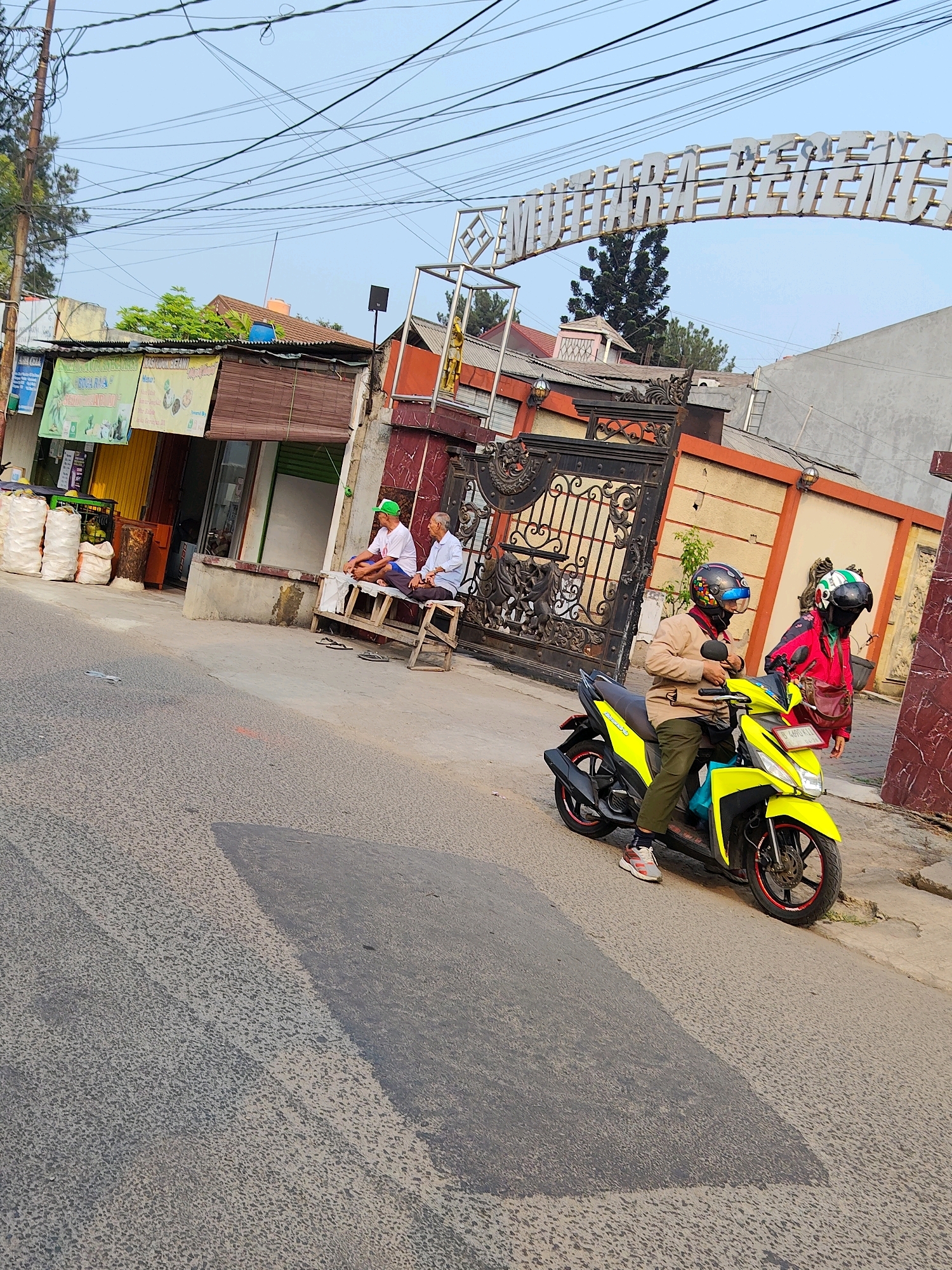 In Bekasi, Indonesia, two men are seated on a wooden bench in front of a building, one wearing a green cap. To their right, a bright yellow scooter with two riders in helmets is parked on the street. The scooter has an Indonesian license plate. Behind the scooter and bench is a large, ornate black gate. Above the gate, a sign arches across, with the text "MUTIARA RESIDENCE" visible. The scene is set on a street with overhead power lines and a clear sky. The buildings in the background are a mix of residential and commercial structures.