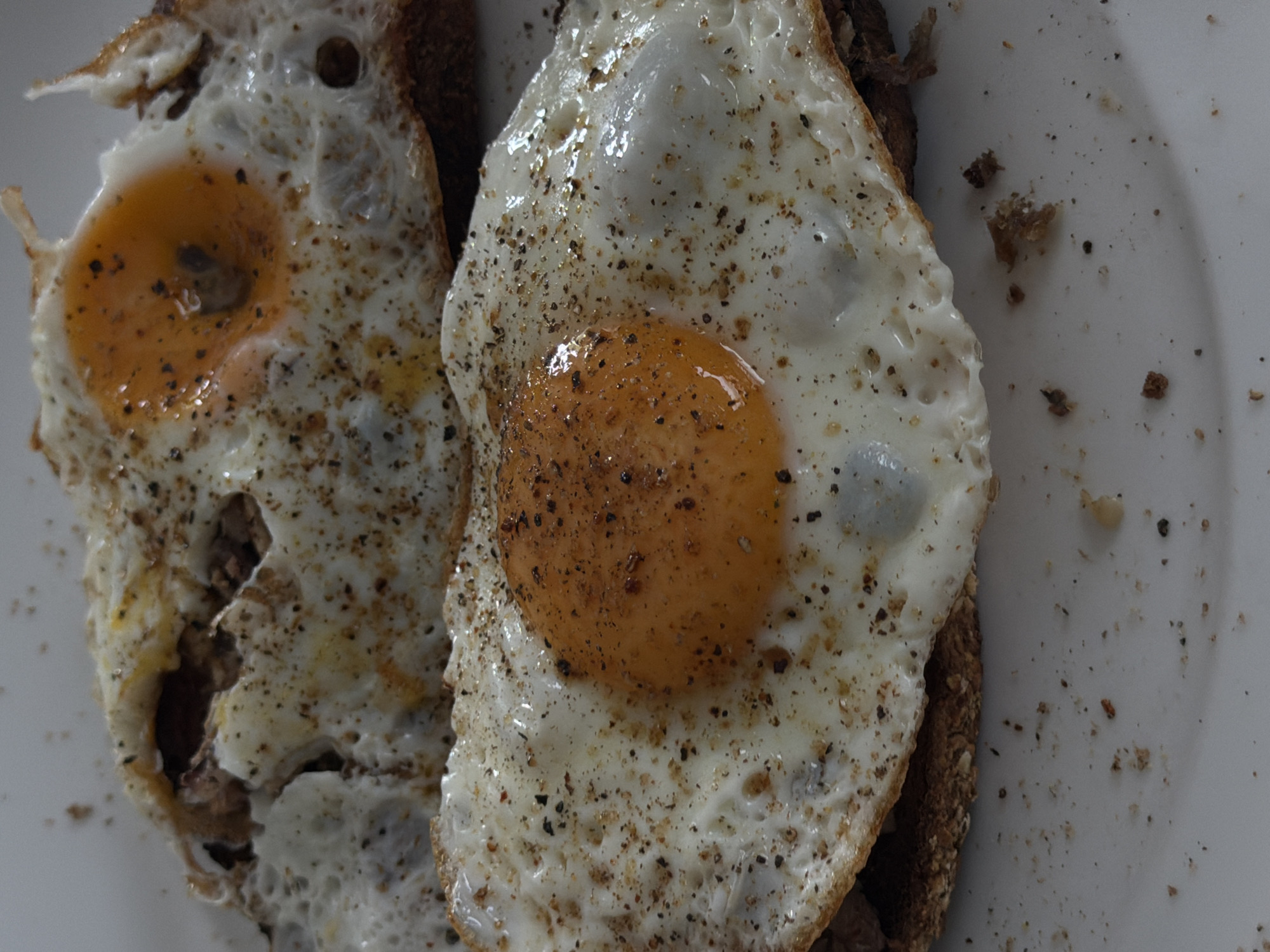 Two fried eggs, seasoned with black pepper, are served on slices of toast. The eggs are golden with slightly crispy edges and the yolks are still soft and runny. The toast appears to be whole wheat. The food is presented on a white plate, which is slightly messy with crumbs. The image is a close-up, overhead shot, with the focus on the food. There are no people visible in the image, and no identifiable landmarks or context to indicate a specific location in London, United Kingdom. The lighting is natural, suggesting it was taken indoors during the day.