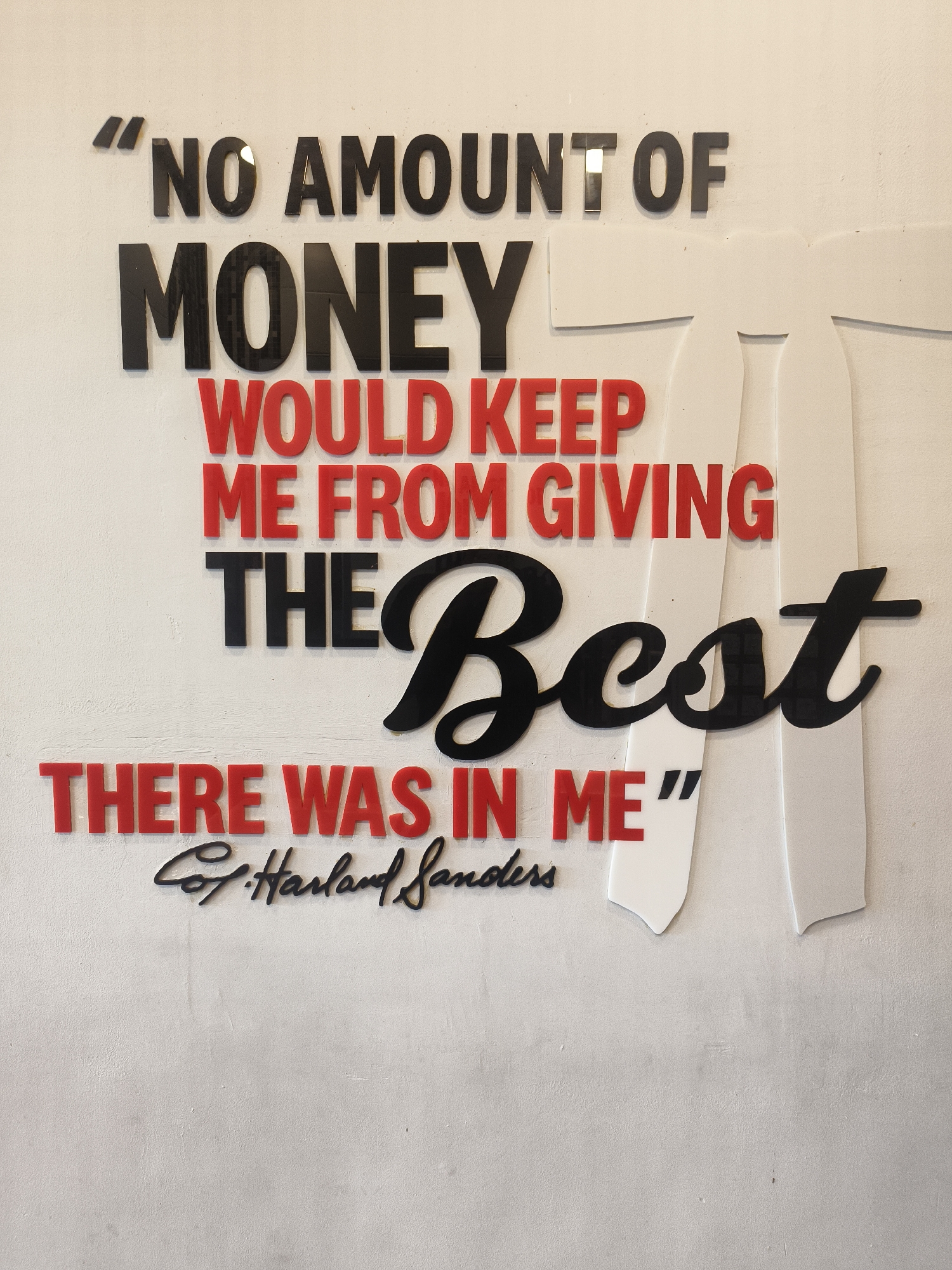 A motivational quote attributed to Col. Harland Sanders is displayed on a white textured wall. The quote reads, "NO AMOUNT OF MONEY WOULD KEEP ME FROM GIVING THE BEST THERE WAS IN ME." The words are presented in three-dimensional block letters. "NO AMOUNT OF" and "MONEY" are in black, while "WOULD KEEP ME FROM GIVING THE" and "THERE WAS IN ME" are in red. The word "Best" is prominently displayed in a large, stylized black font. Two white cutouts resembling bowties are positioned behind the text, one on the right and one slightly overlapping the center. The signature "Col. Harland Sanders" is inscribed in cursive black letters below the main quote. The overall presentation is clean and modern, suggesting it might be a decorative element in a restaurant or a themed establishment. The context of the location in Denpasar, Indonesia, is not discernible from the visual information provided.