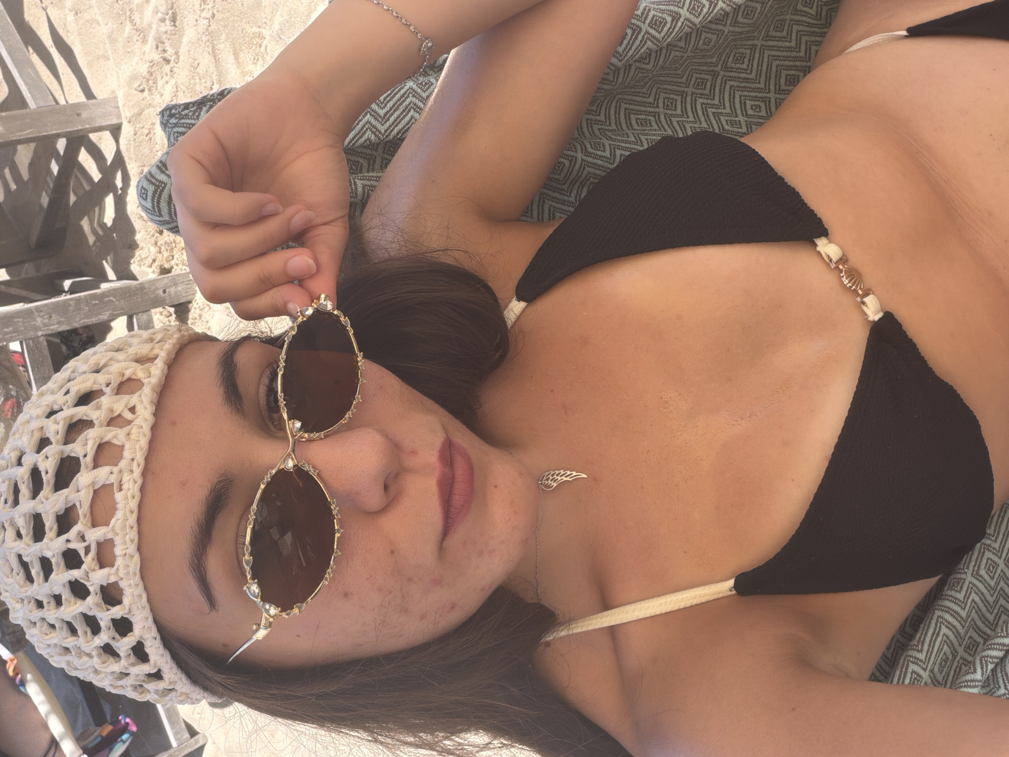 A young woman is lying on a patterned blanket on a sandy beach, with wooden beach chairs in the background. She is wearing a white crocheted hat, gold-rimmed sunglasses with decorative embellishments, and a black bikini. A silver necklace with a wing pendant is visible around her neck. Her right hand is resting near her face, adjusting her sunglasses. The lighting suggests it is daytime, and the overall scene depicts a relaxed beach setting.